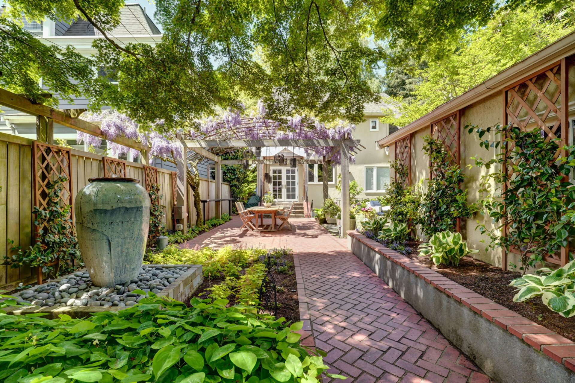 A peaceful backyard garden with lush greenery, a pergola adorned with wisteria, and a large decorative pot along a brick pathway leading to a house.