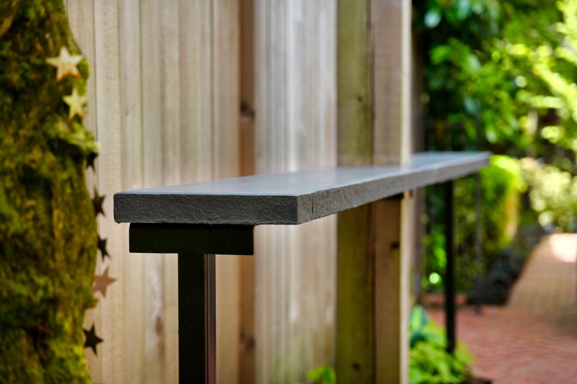 A sleek, narrow shelf is mounted on a wooden fence beside a lush, green garden path with a peaceful, natural atmosphere.