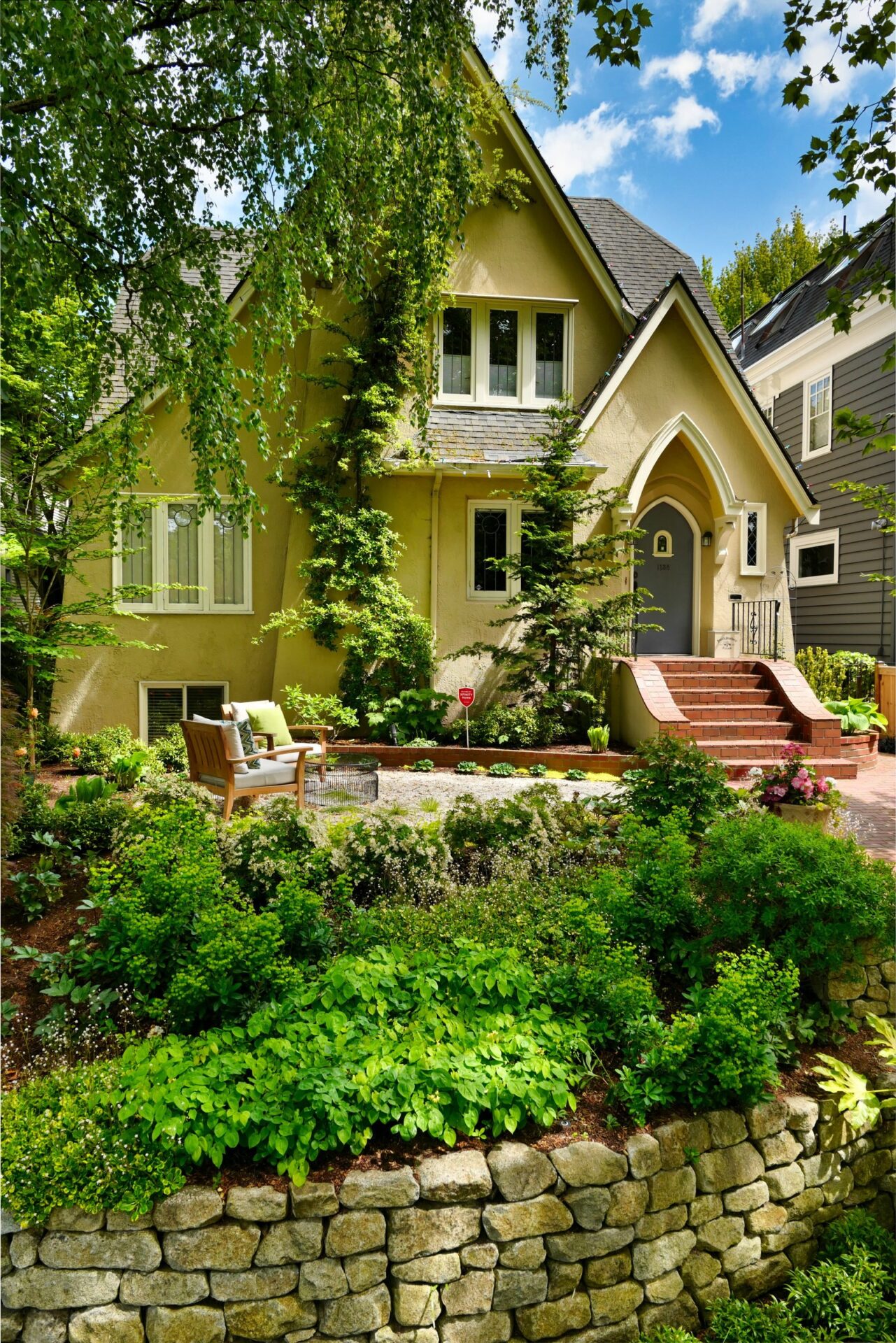 Charming house with lush garden, stone wall, and patio seating. Gabled roof and arched entrance enhance its picturesque, cottage-style appearance amidst greenery.