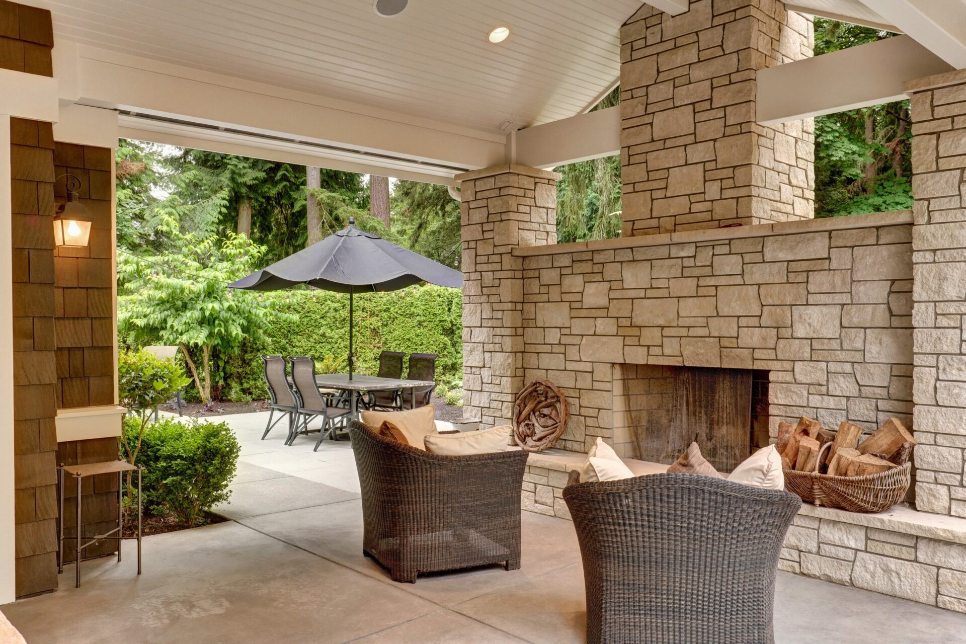 Covered patio with stone fireplace, wicker chairs, and outdoor dining set. Surrounded by lush greenery and trees. Bright, airy atmosphere without people.