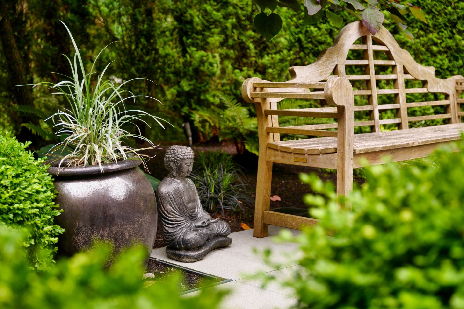 A serene garden featuring a wooden bench, a Buddha statue, and a large potted plant surrounded by lush greenery creates a peaceful ambiance.