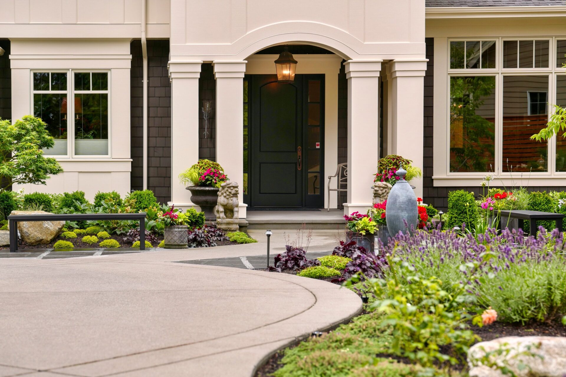 Elegant house entrance with a black door, surrounded by well-maintained gardens, colorful flowers, and clean pathways. Inviting and picturesque residential scene.