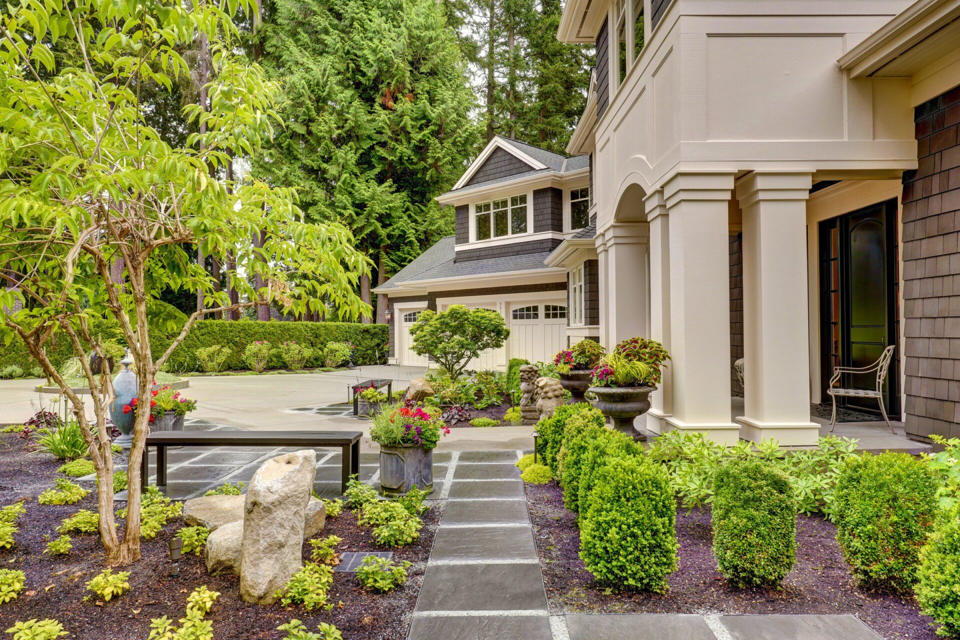 Modern house with landscaped garden, featuring neatly trimmed hedges, potted plants, and a small seating area. A person walks along the sidewalk.