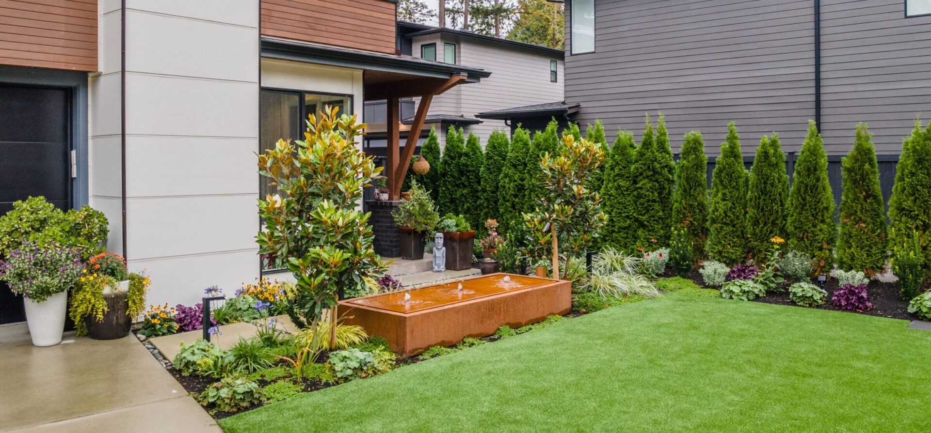 A modern garden with neatly trimmed hedges, flowering plants, and a central water feature, nestled between contemporary houses with wooden and gray exteriors.