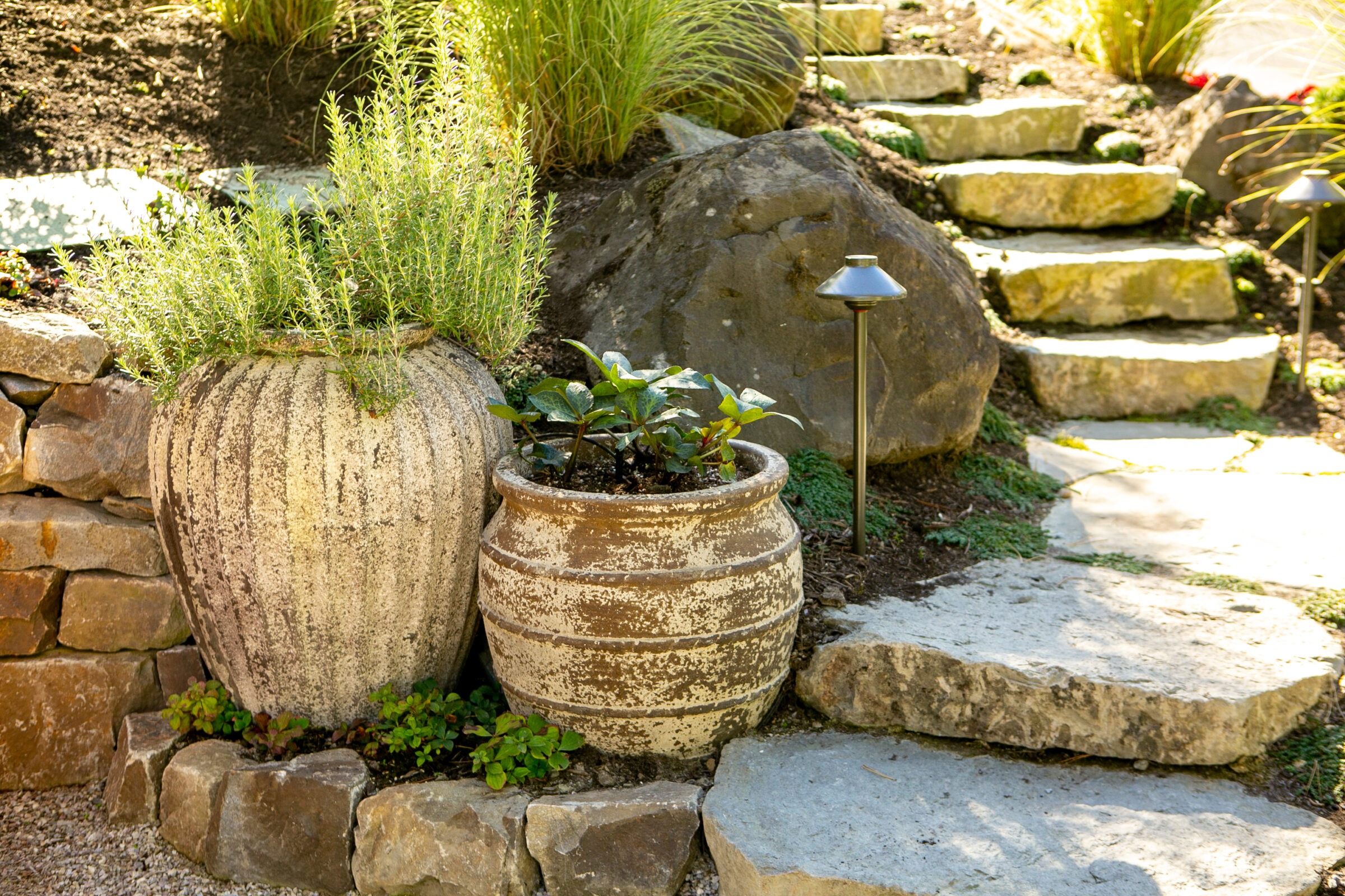 Stone steps lead through a garden with large planters and greenery. A small pathway light illuminates the natural, rustic setting.