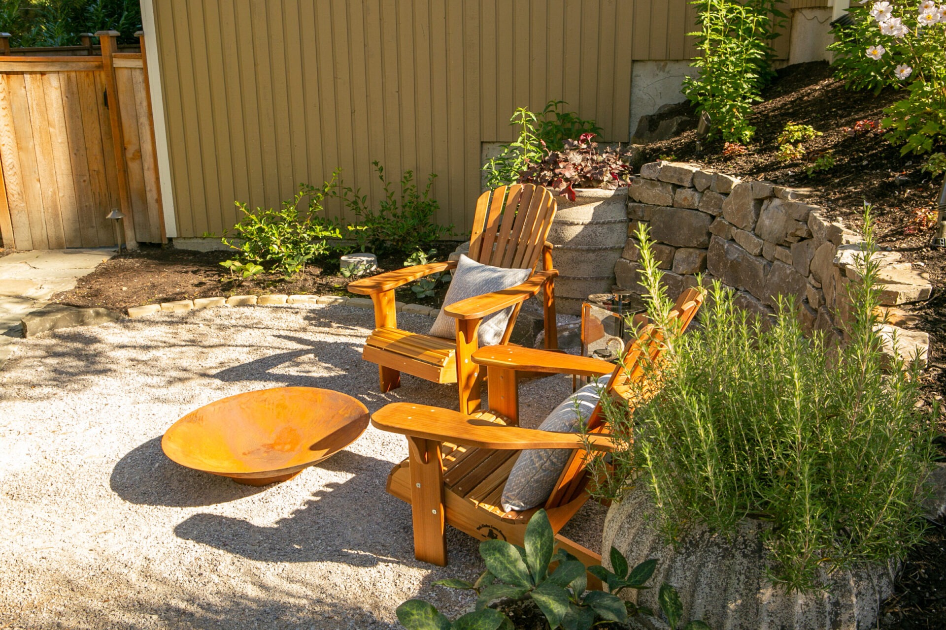 Sunny garden patio with two wooden chairs, a round fire pit, and lush greenery. Stone wall and wooden fence provide a cozy setting.