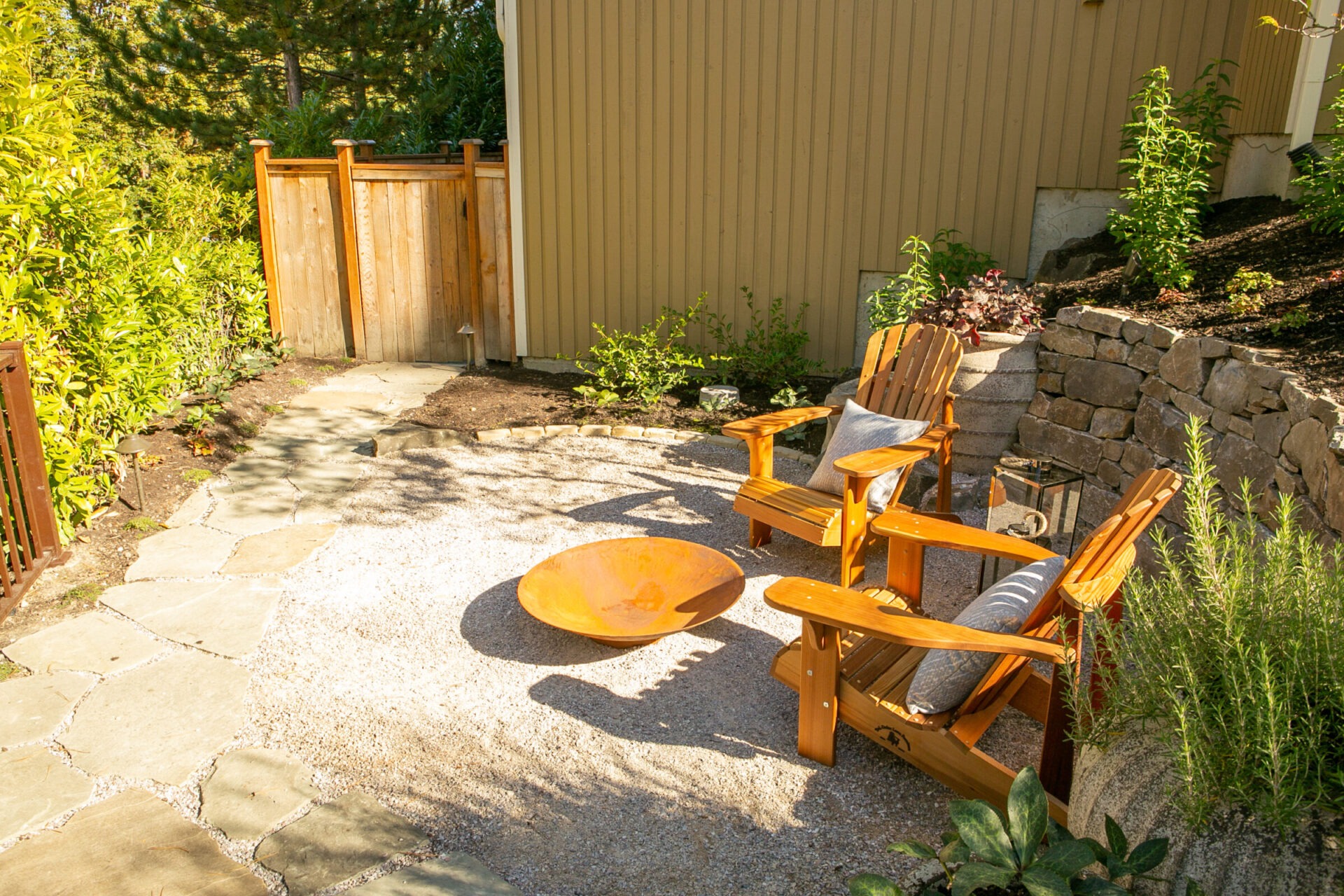 A sunny backyard with two wooden chairs, a fire pit, stone pathway, and lush greenery surrounded by a wooden fence and beige structure.