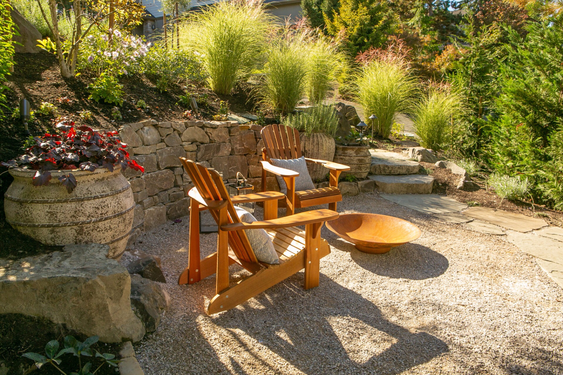 Cozy outdoor seating area with two wooden chairs, a fire pit on gravel, surrounded by lush greenery and stone walls. Peaceful garden setting.