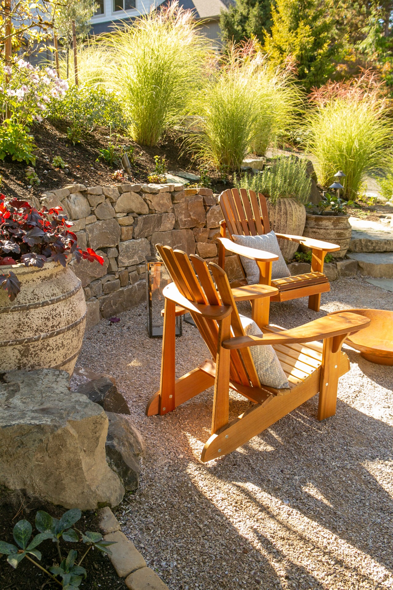 A serene garden scene with wooden chairs, a stone wall, vibrant plants, and potted flowers under sunlight, creating a peaceful outdoor retreat.