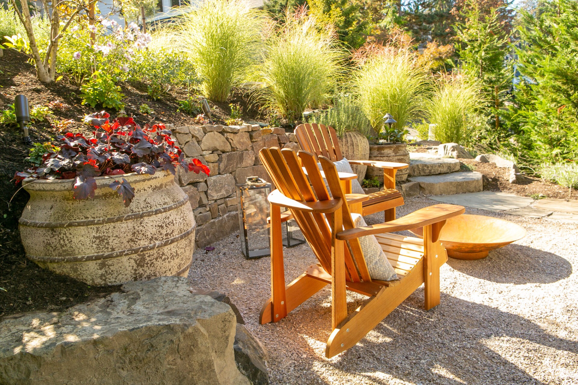 A serene garden scene with wooden chairs, a large stone planter, vibrant flowers, and abundant greenery under dappled sunlight, creating a relaxing atmosphere.