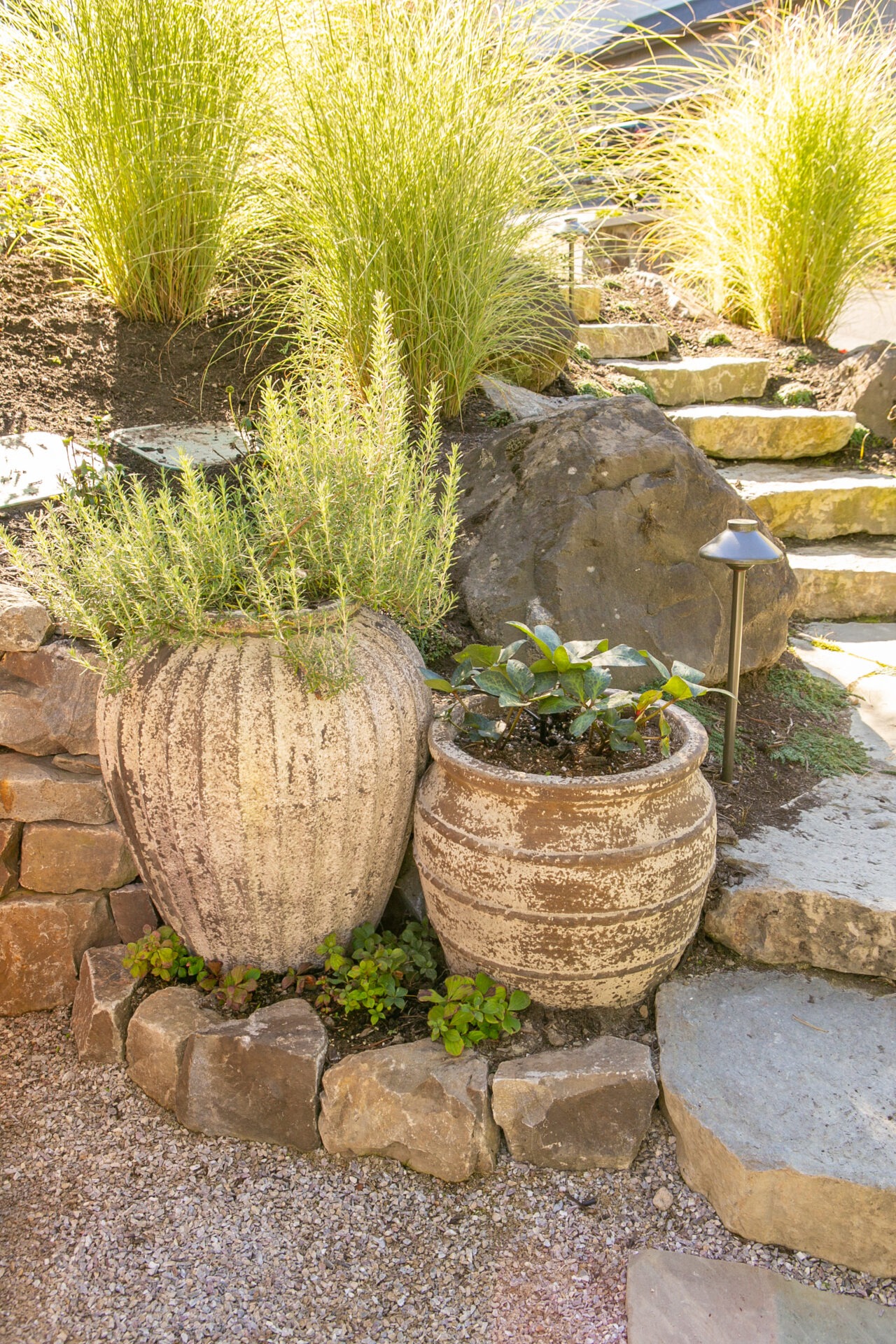 Stone planters with green plants sit on a gravel path, surrounded by rocks and grass, near stone steps under sunlight.