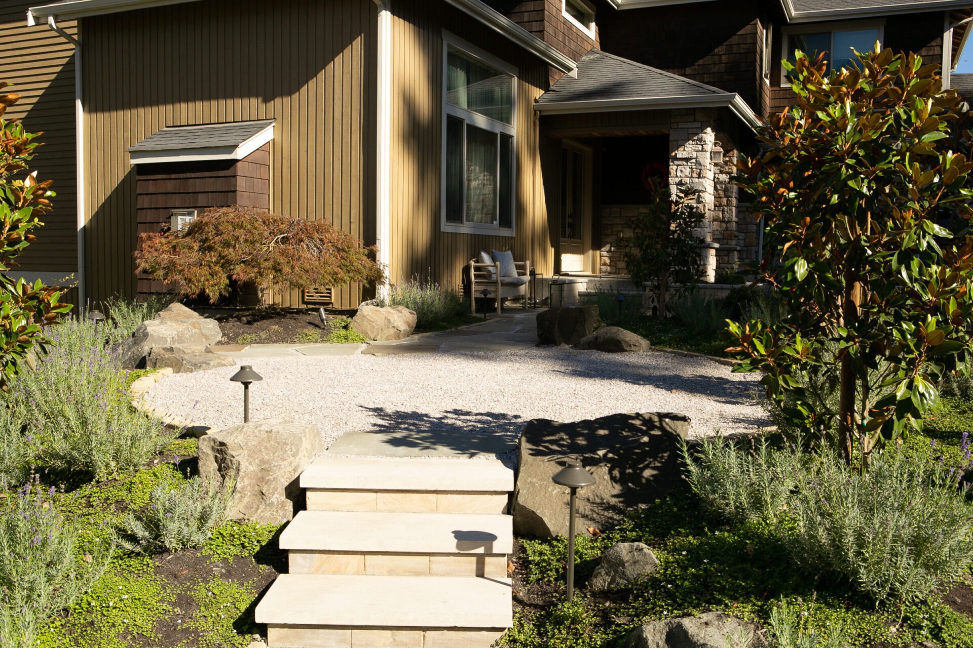 Modern house entrance with greenery, stone steps, and gravel path. Sunlit facade and small garden create a welcoming, natural atmosphere. No people visible.