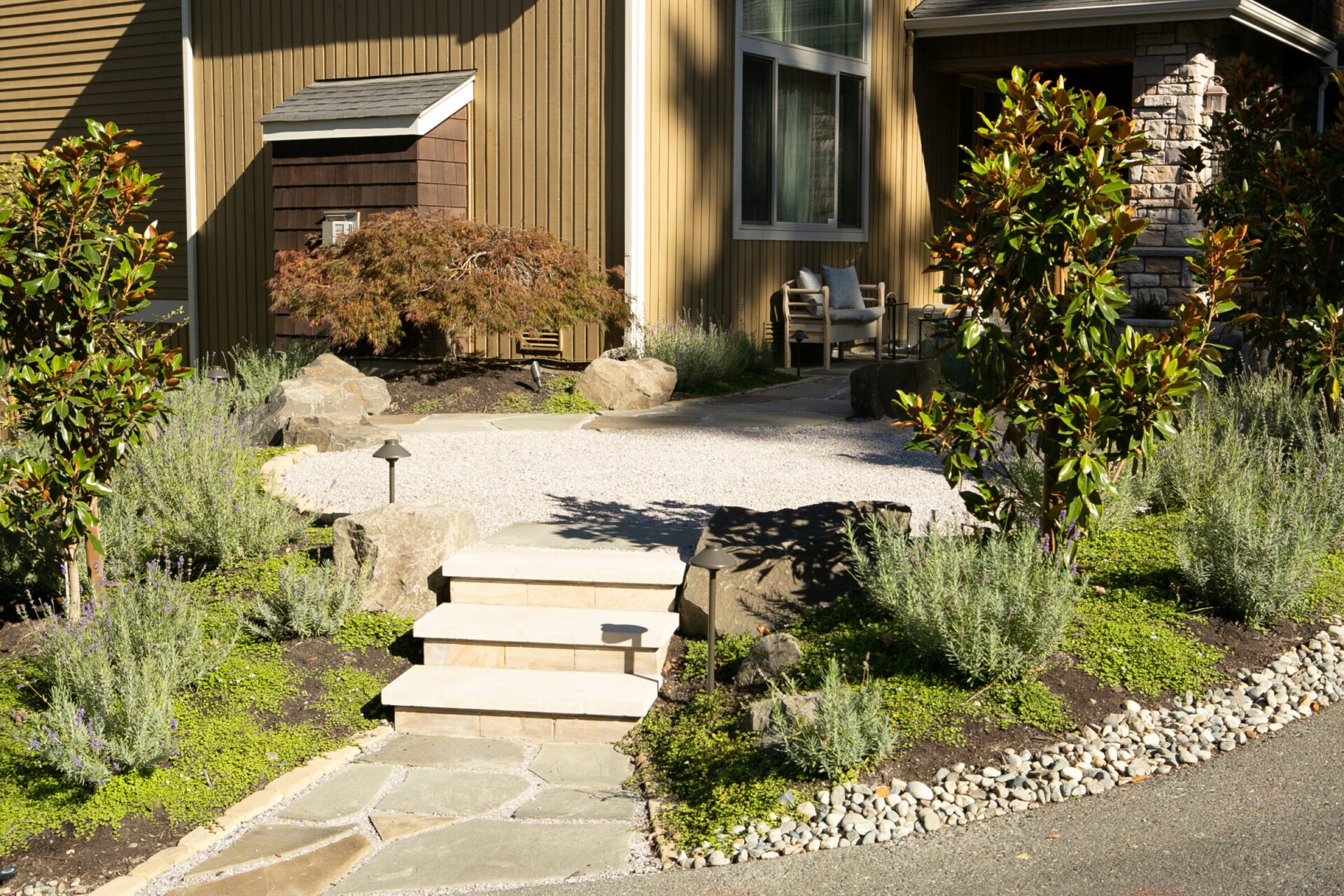 A landscaped walkway with stone steps, shrubs, and a modern house exterior featuring large windows and light brown siding, surrounded by greenery.