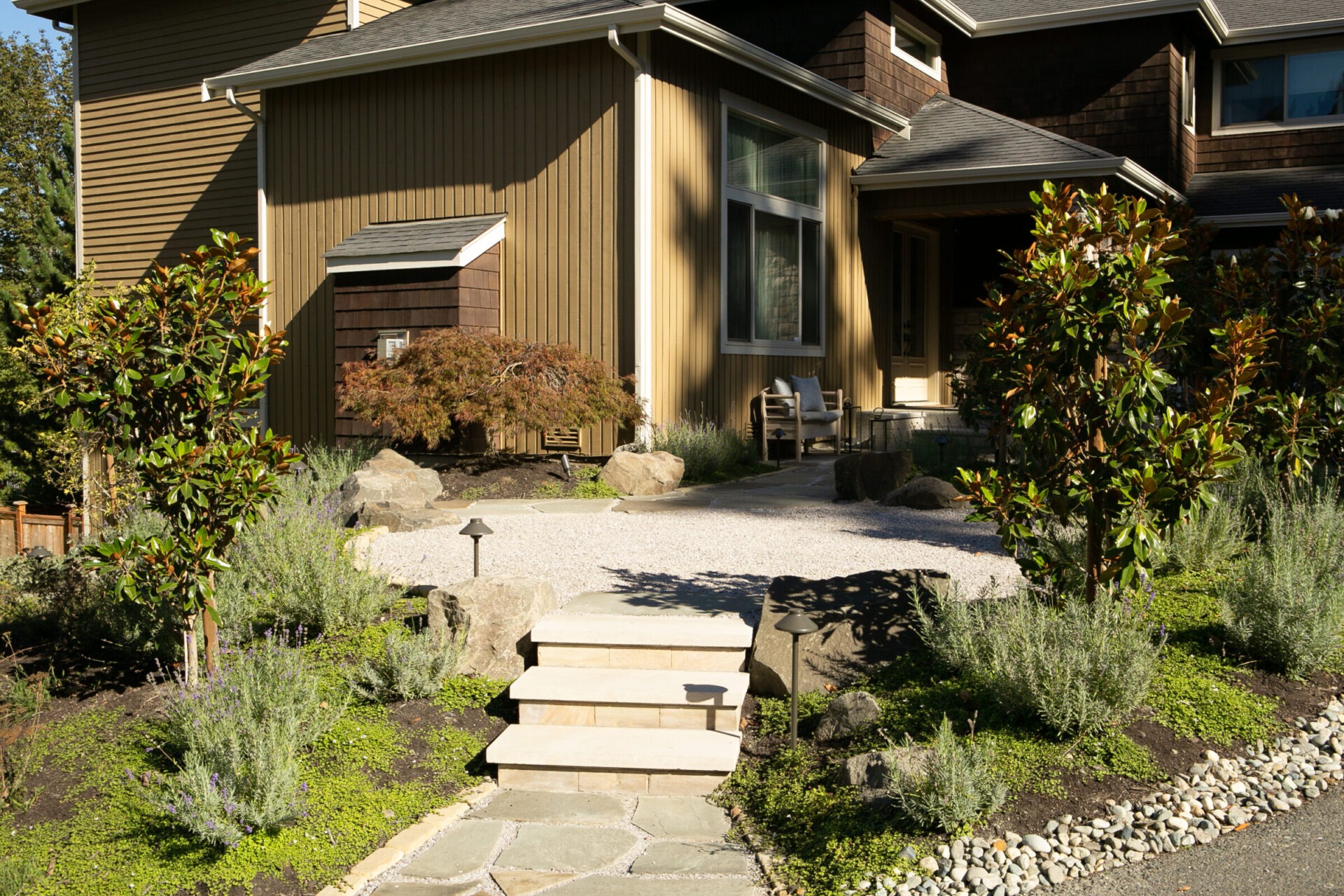 A modern house with a neatly landscaped front yard, featuring stone steps, greenery, and decorative rocks under a clear blue sky.