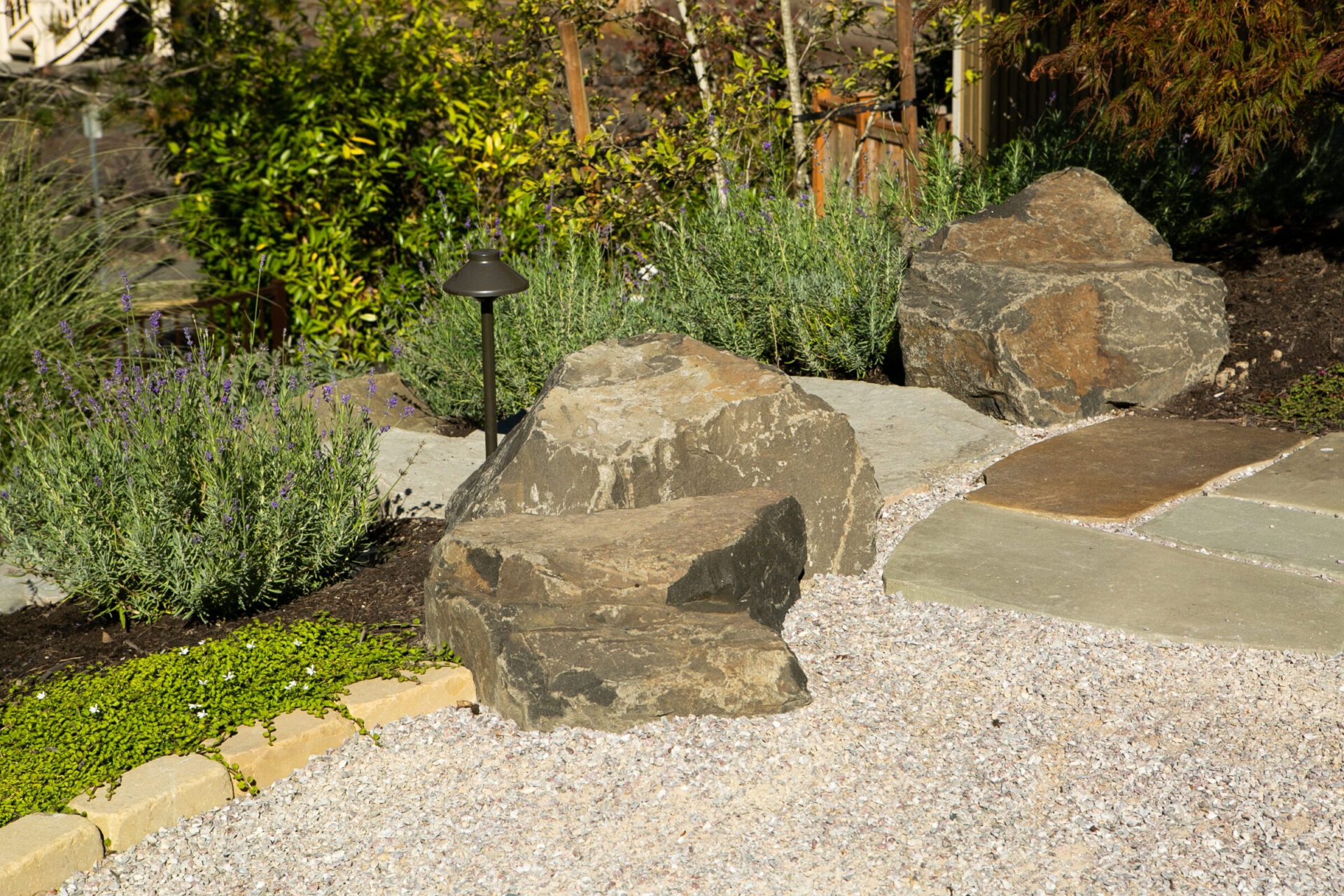 A serene garden scene features large rocks, greenery, and a gravel path, accented by a small garden light and lush shrubs.