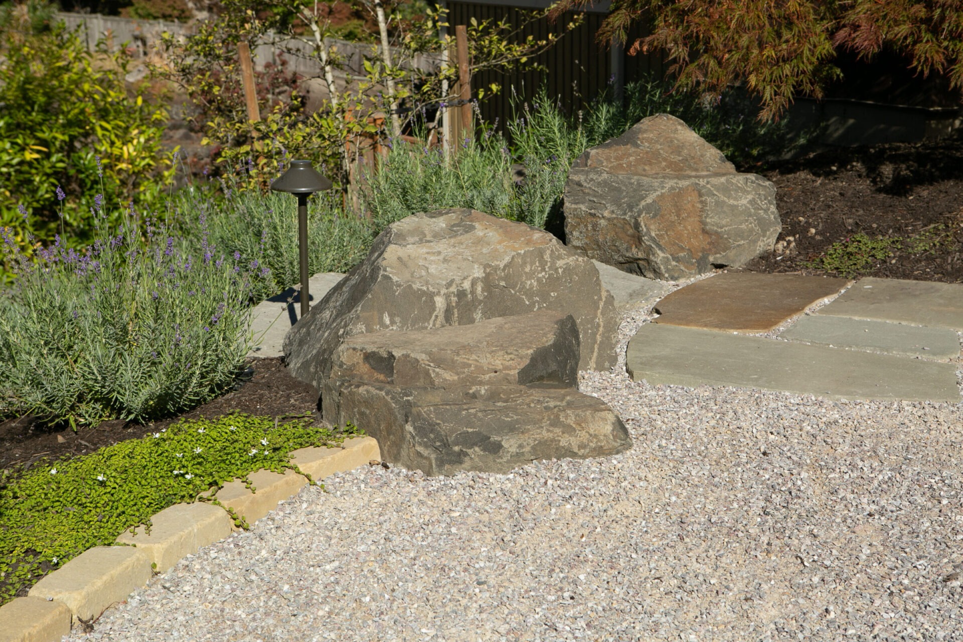 A garden features large rocks, gravel pathway, greenery, and a small lamppost, creating a serene, natural outdoor setting.