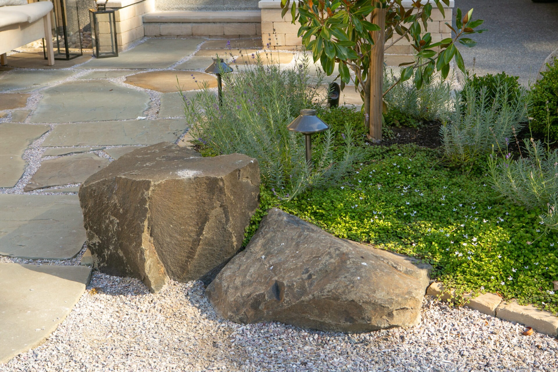 A serene garden setting with two large rocks, green plants, gravel, and a pathway. Nearby, small garden lights add charm.