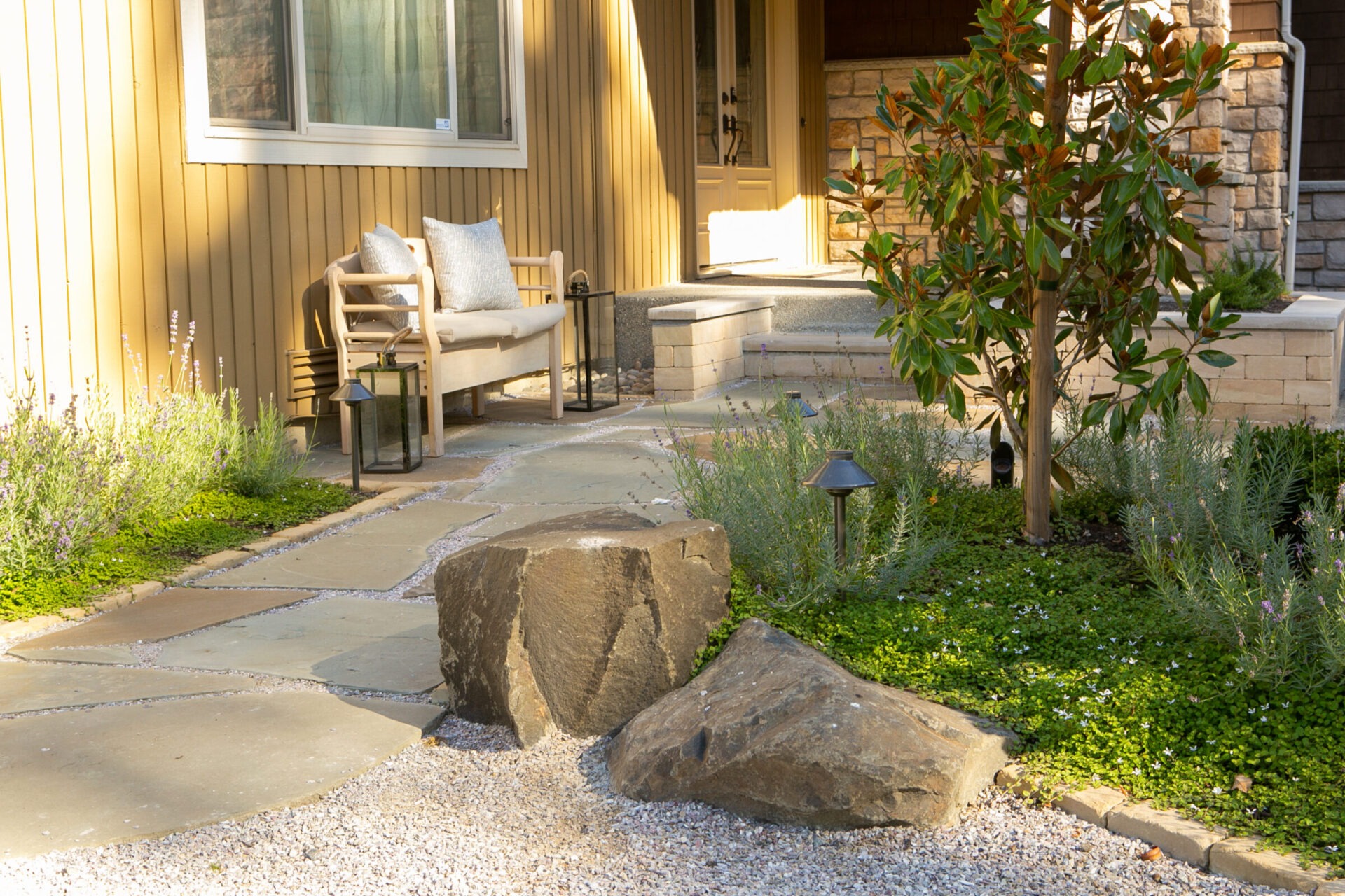 A cozy patio with bench, plants, and rocks beside a stone pathway leading to a house entrance. Sunlight casts warm shadows.