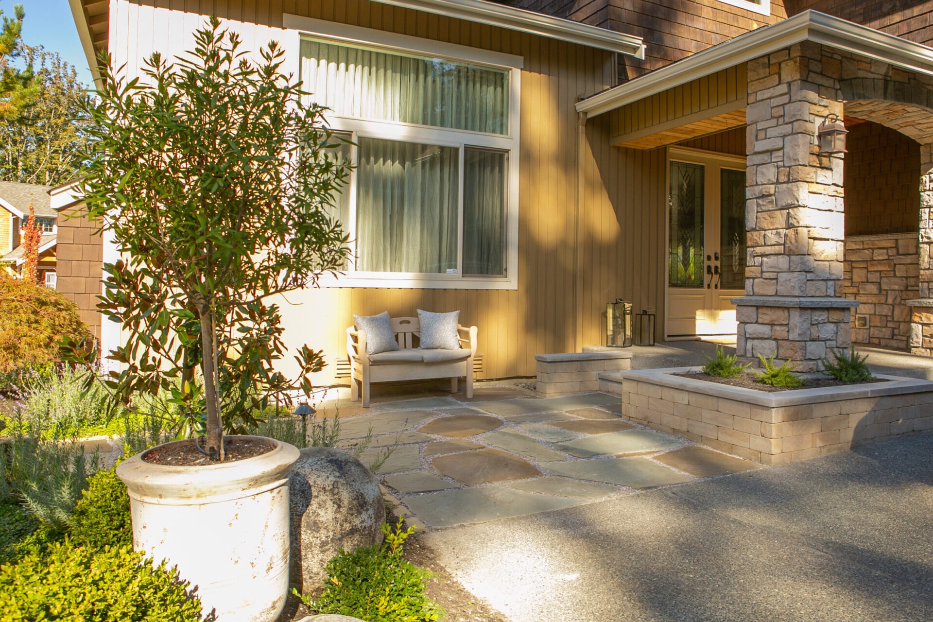 A cozy house entrance with a stone porch, bench, and potted plants. Sunlit and inviting, perfect for a peaceful retreat.