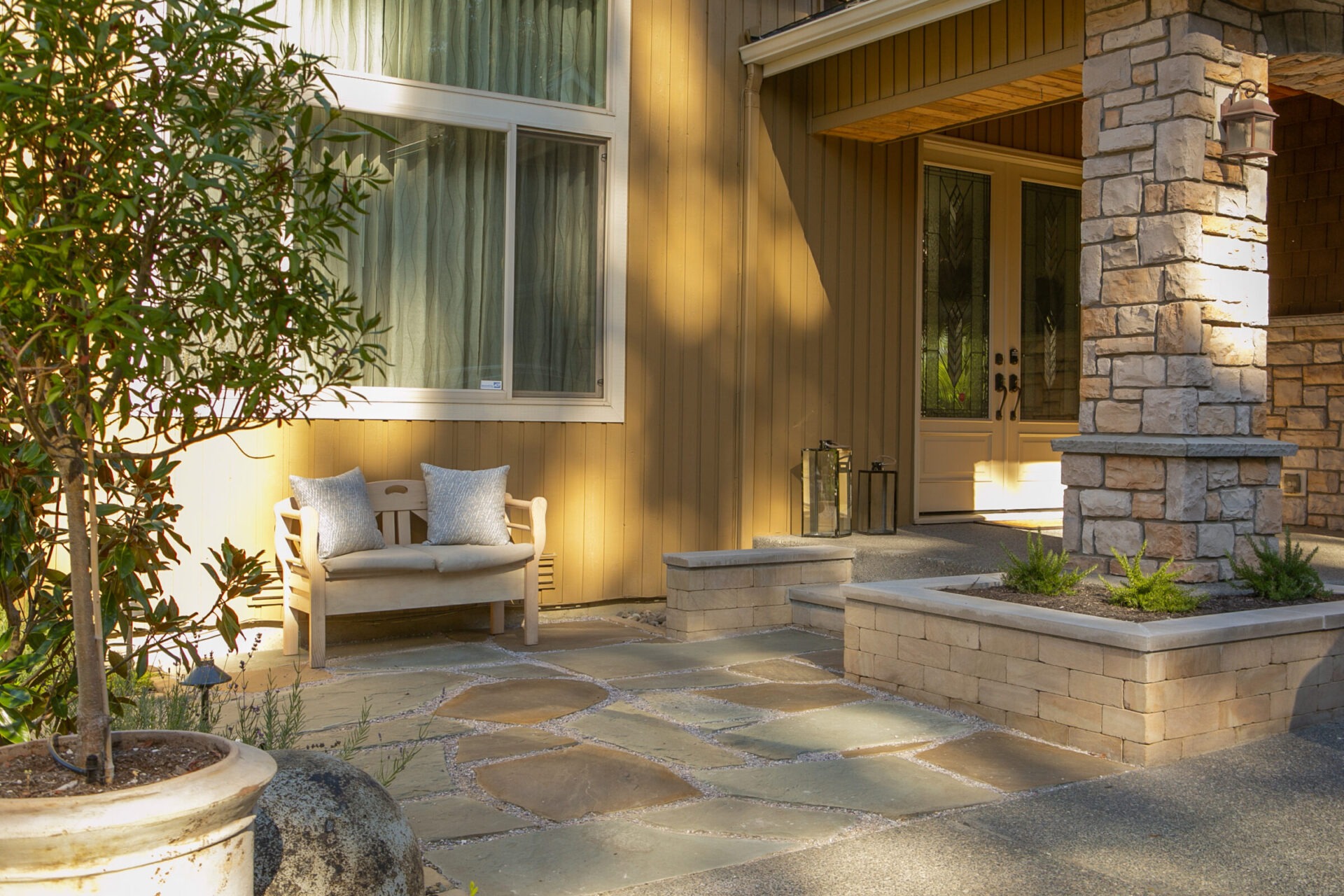 The image shows a peaceful porch with a stone pillar, bench with cushions, and potted plants, casting soft shadows in natural light.