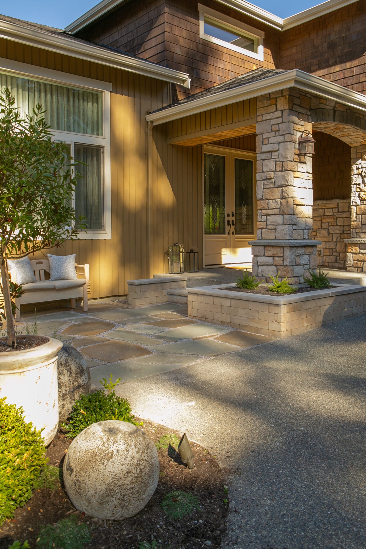 Modern house entrance with stone and wood accents, shaded by trees. A bench and decorative sphere enhance the inviting atmosphere. Sunlight creates warm tones.