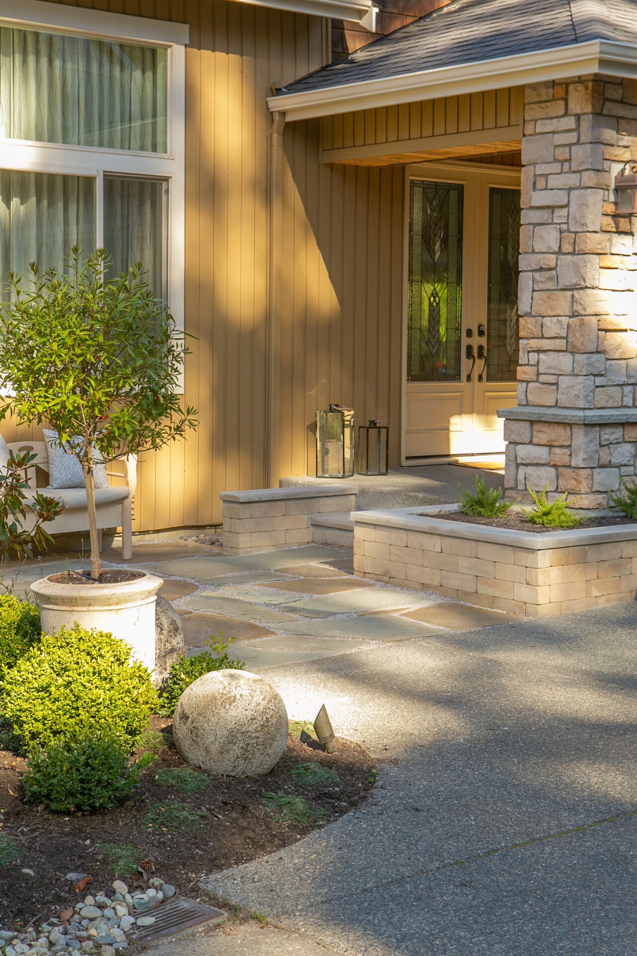 A cozy front entrance featuring stone columns, potted plants, and a well-maintained pathway, bathed in warm sunlight and inviting ambiance.