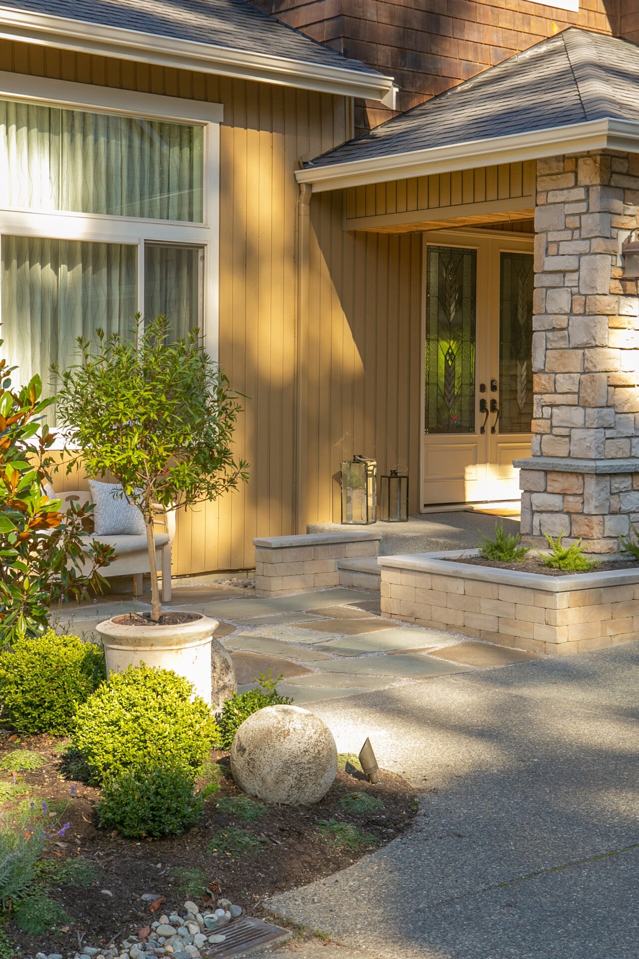 A cozy house entrance with stone pillars, potted plants, and a small garden. Sunlight casts soft shadows on the walls and walkway.