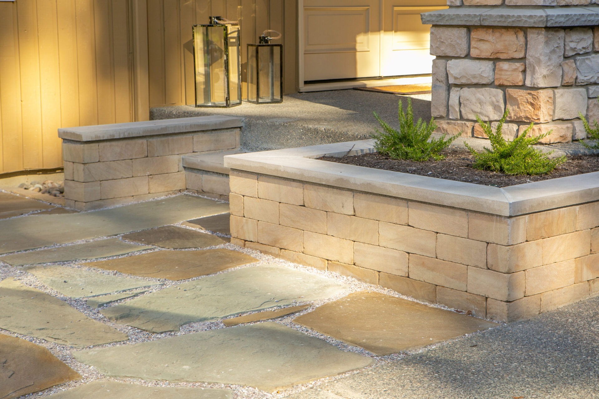 A stone patio with raised planters, two lanterns, and a house entrance. Sunlight casts warm shadows on the textured surface.