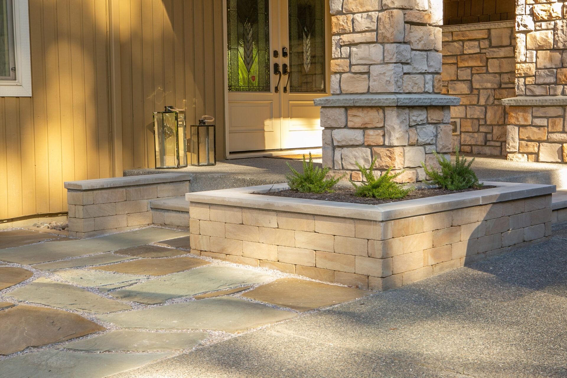 Modern entrance with stone pillars and planter boxes, leading to a wooden double door. Soft lighting highlights the clean, contemporary design.