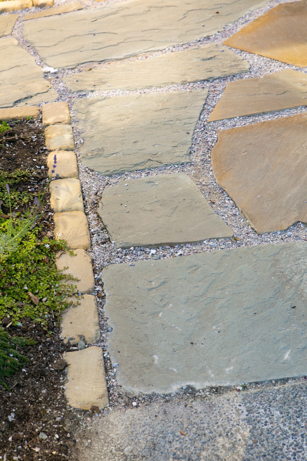 A stone pathway with irregular slabs and gravel accents, bordered by small plants and a garden area, creating a rustic outdoor ambiance.