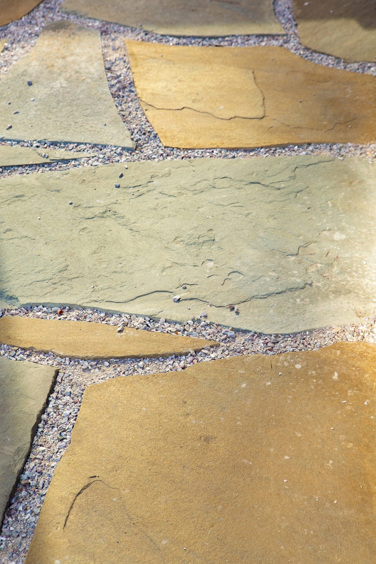 Close-up of a stone pathway with irregular, tan stones and small gravel filling the gaps, under bright sunlight, creating a textured appearance.