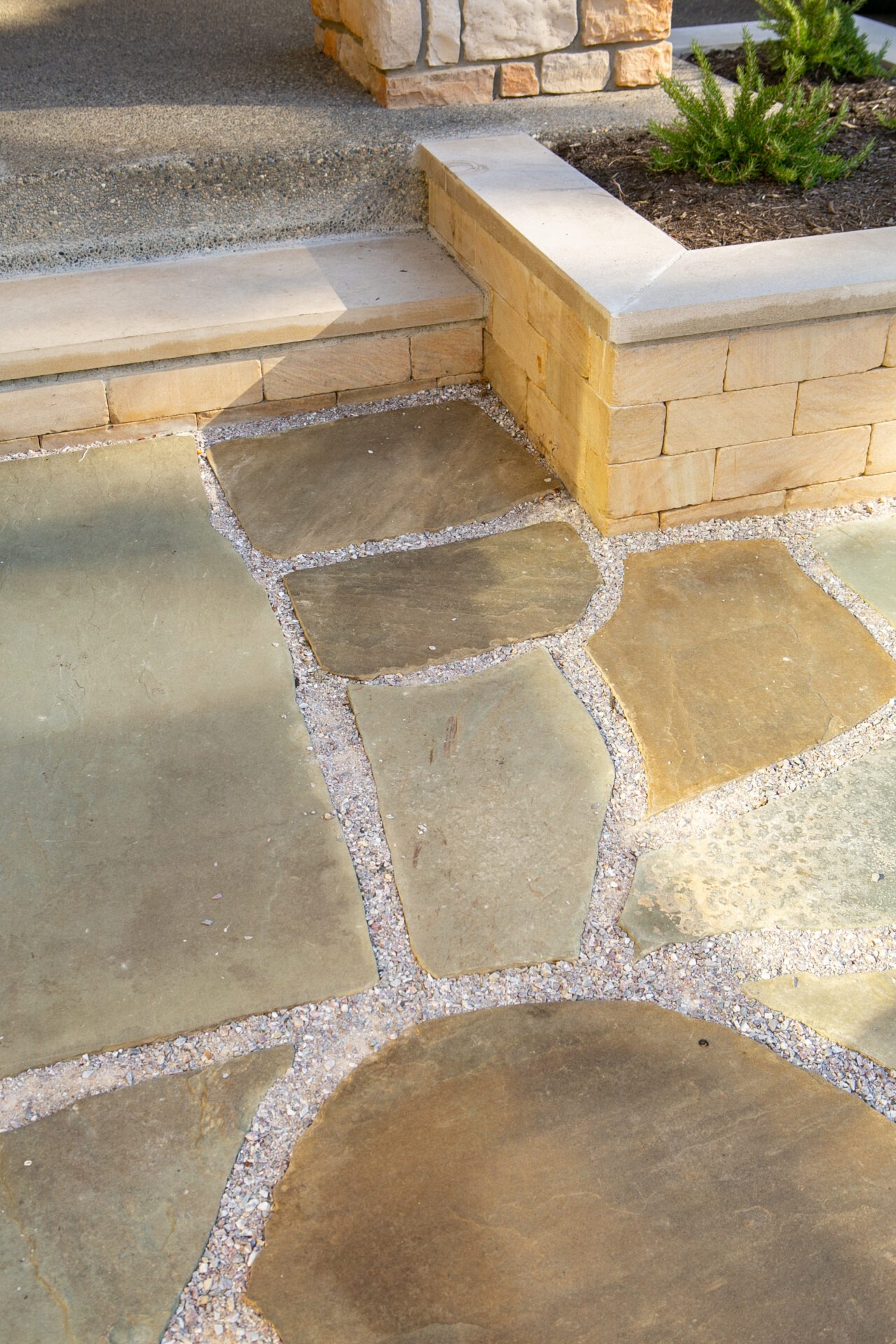 Stone pathway with irregular flagstone pavers leads to a low stone wall and small garden. Sunlight casts soft shadows across the surface.