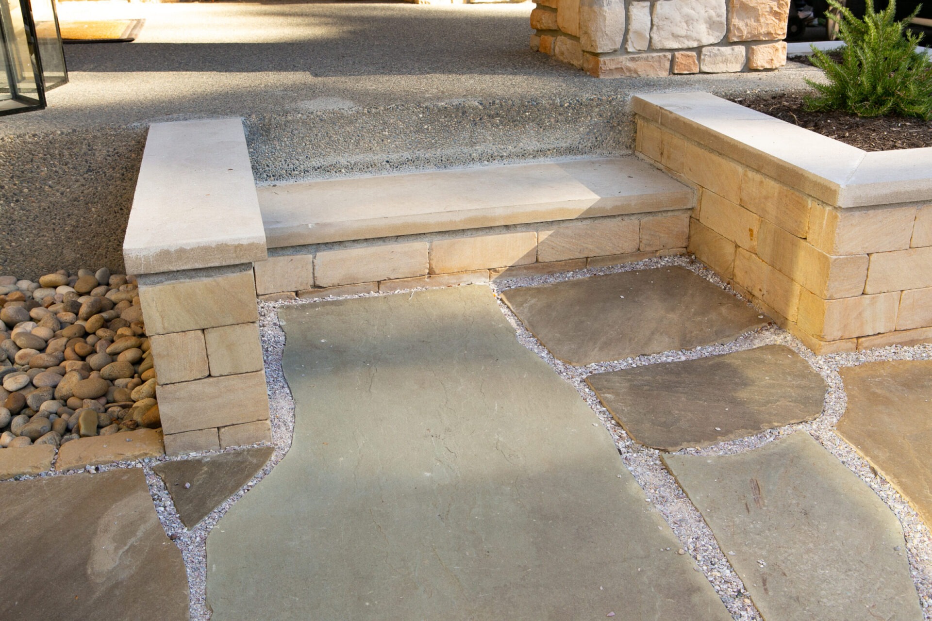 Stone patio with beige bricks and gravel accents, featuring a small step. Pebbles and a plant border the space, creating a neat appearance.