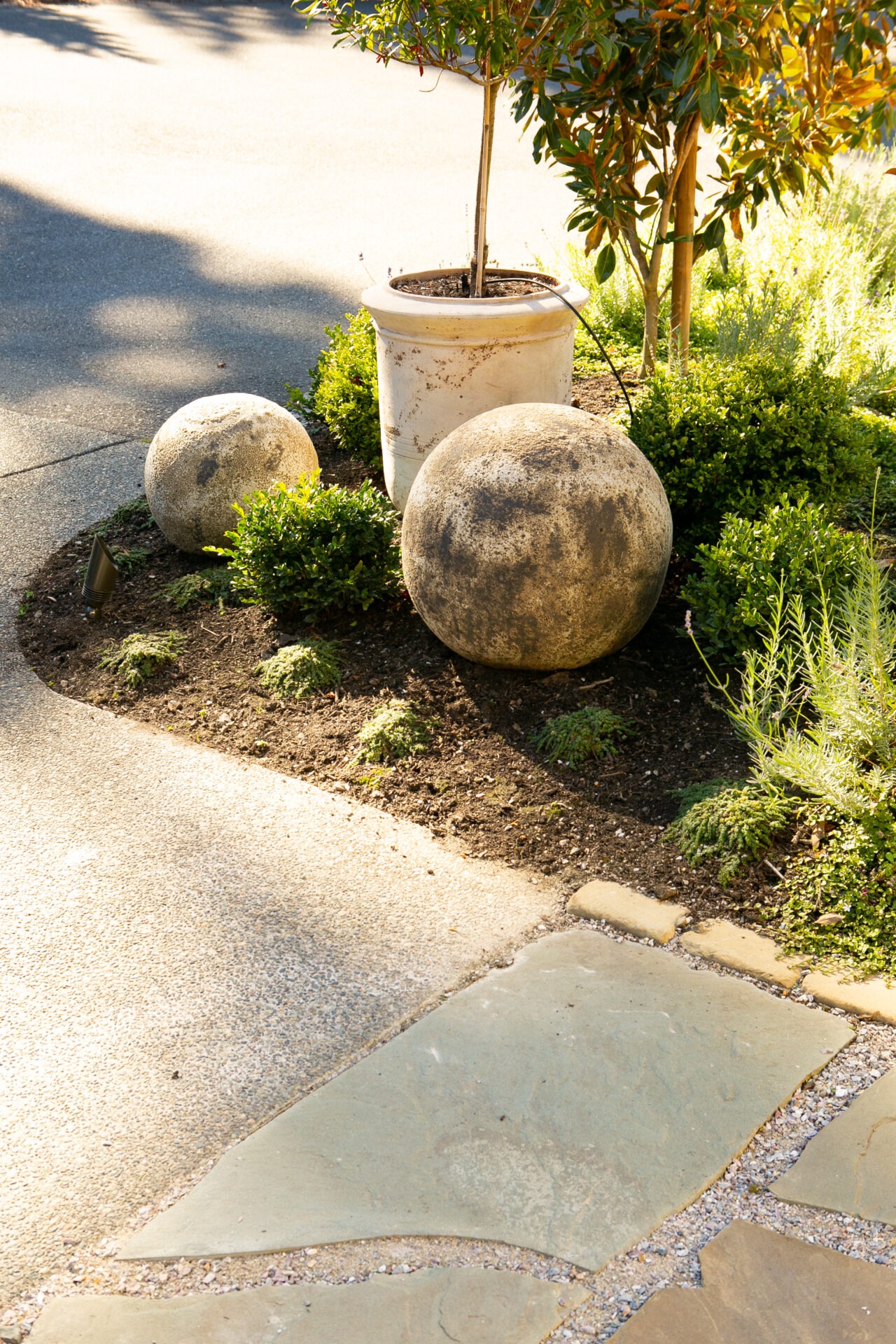 A garden scene with spherical stone sculptures and a potted plant surrounded by greenery on a sunny day, near a stone walkway.