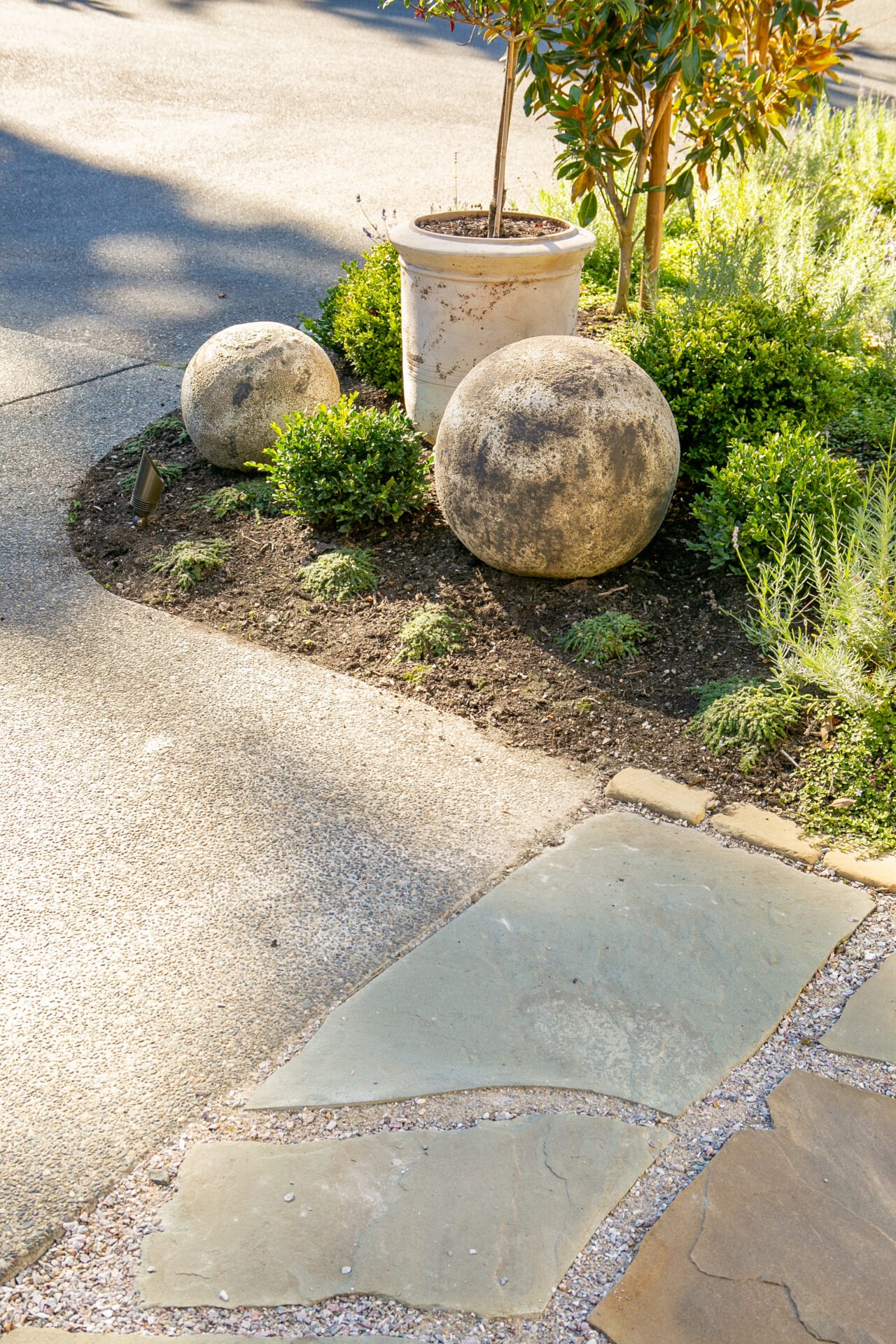 A garden scene with two stone spheres beside a planter. Surrounded by greenery, pathways feature both stone and pavement surfaces.