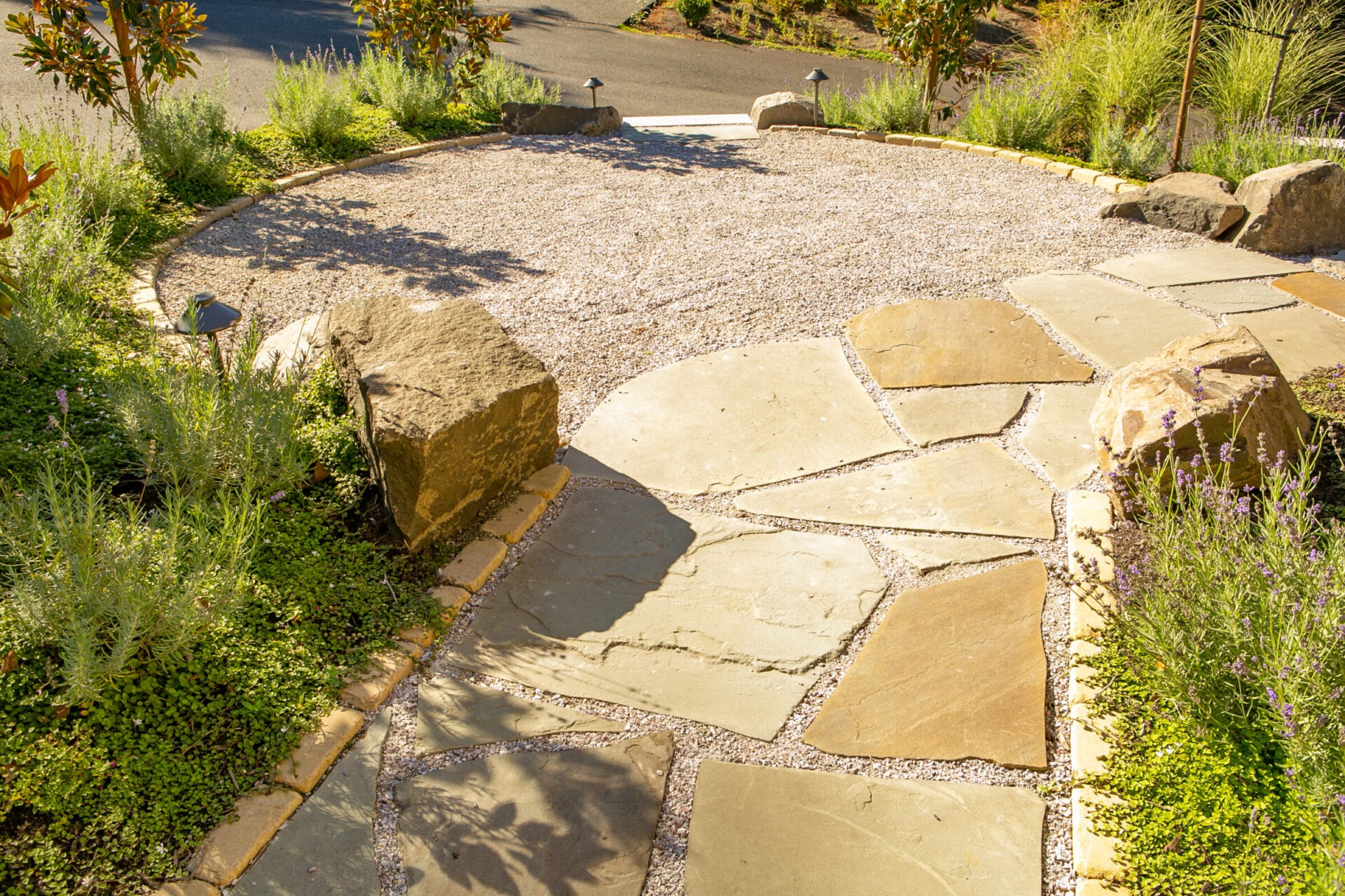 A sunlit garden path with large stones and gravel, surrounded by greenery, leads to a circular area with decorative rocks.