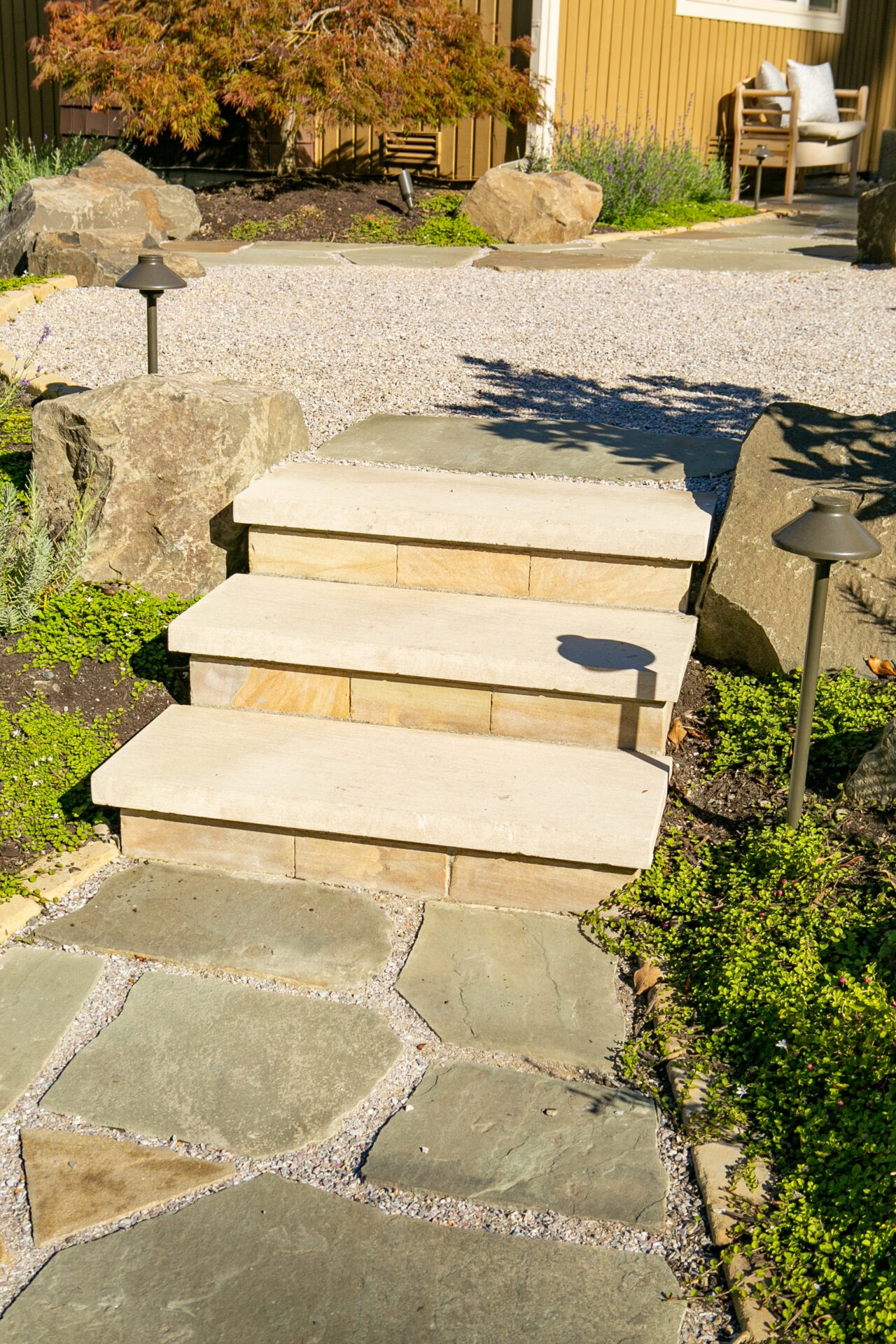 Stone steps and pathway surrounded by greenery and shrubs, leading to a gravel area with a chair nearby, lit by garden lamps.