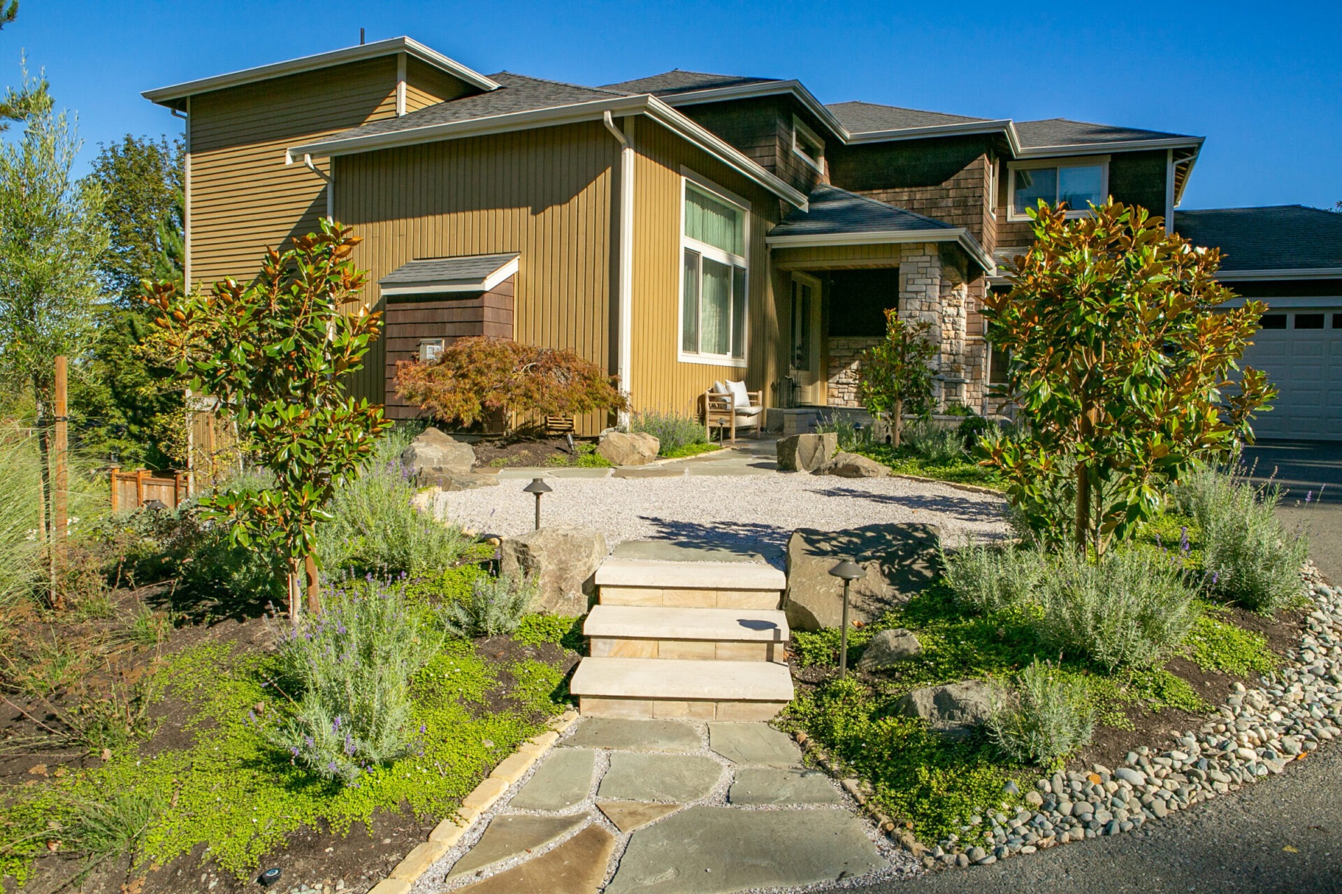 Modern two-story house with landscaped garden, stone path, and wooden exterior. Surrounded by greenery under a clear blue sky. Quiet suburban setting.