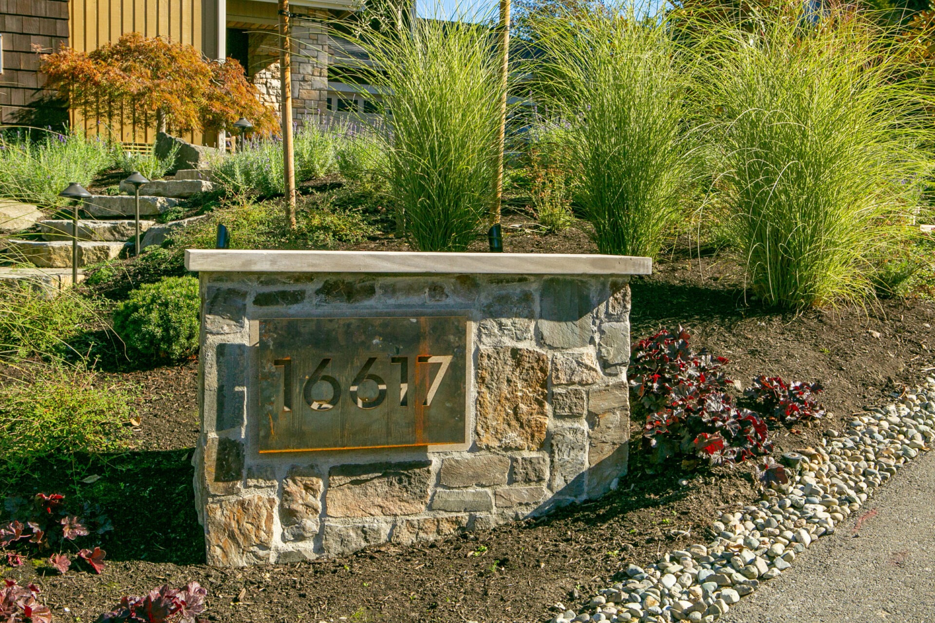 Stone address marker with number 16617 set in a landscaped garden with ornamental grasses and shrubs, adjacent to a modern building.