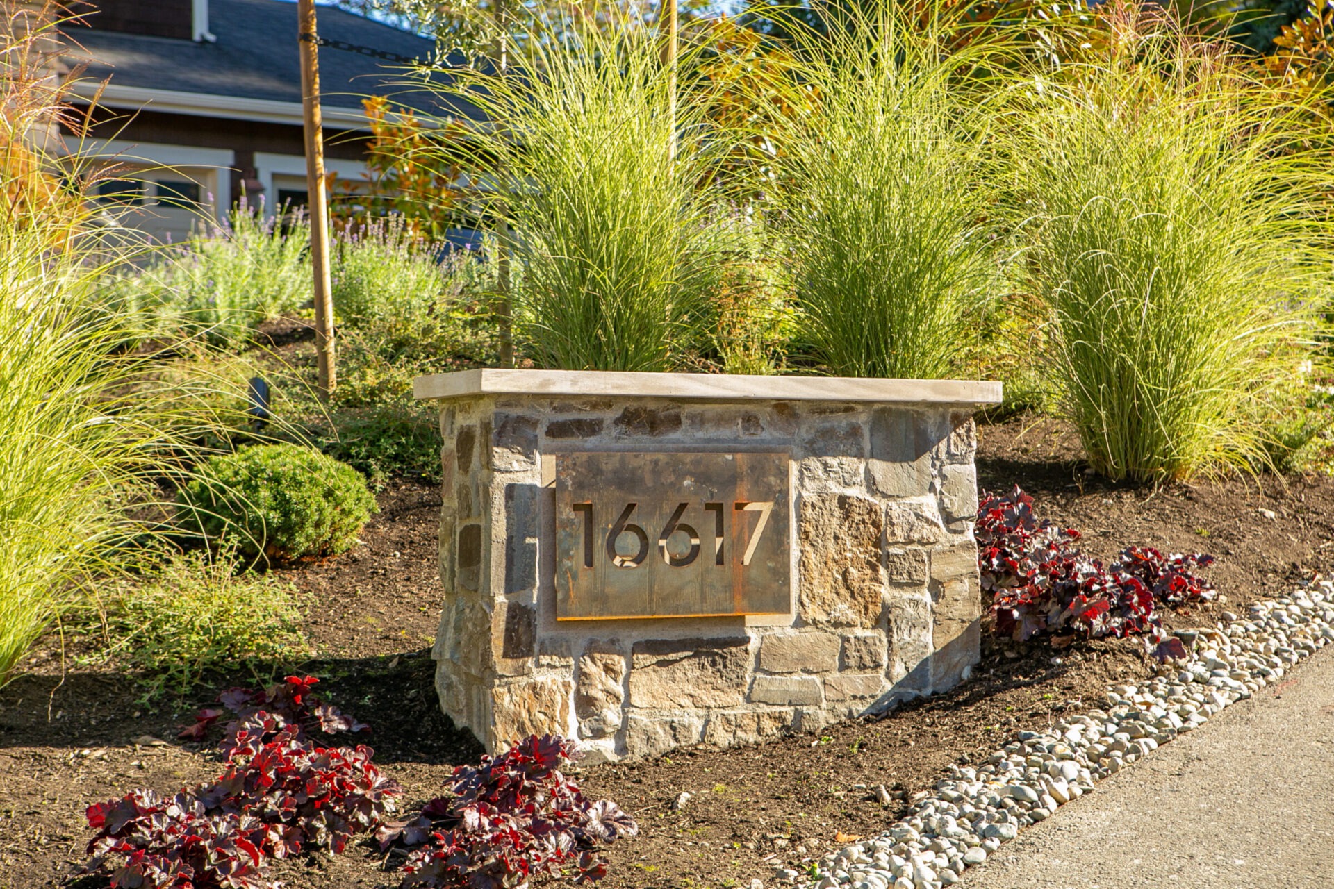 A stone pillar with the number 16617 is surrounded by plants and greenery, situated outdoors near a building in daylight.