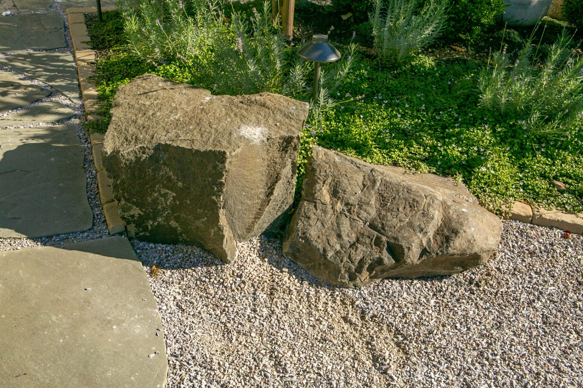 Two large rocks on a gravel surface, surrounded by greenery and a small garden light. Sunlit, outdoor setting with natural landscaping elements.