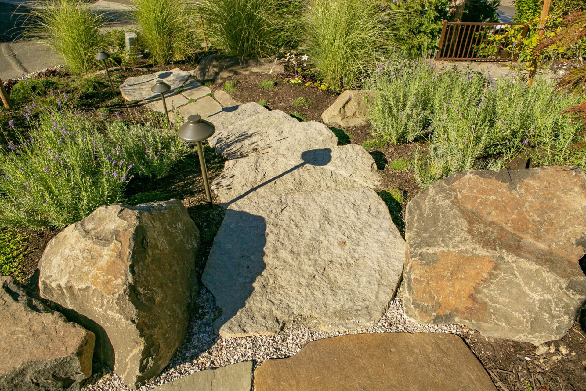 Stone pathway with large rocks, surrounded by lush greenery and garden lighting, under bright sunlight. No recognizable landmarks or historical buildings present.