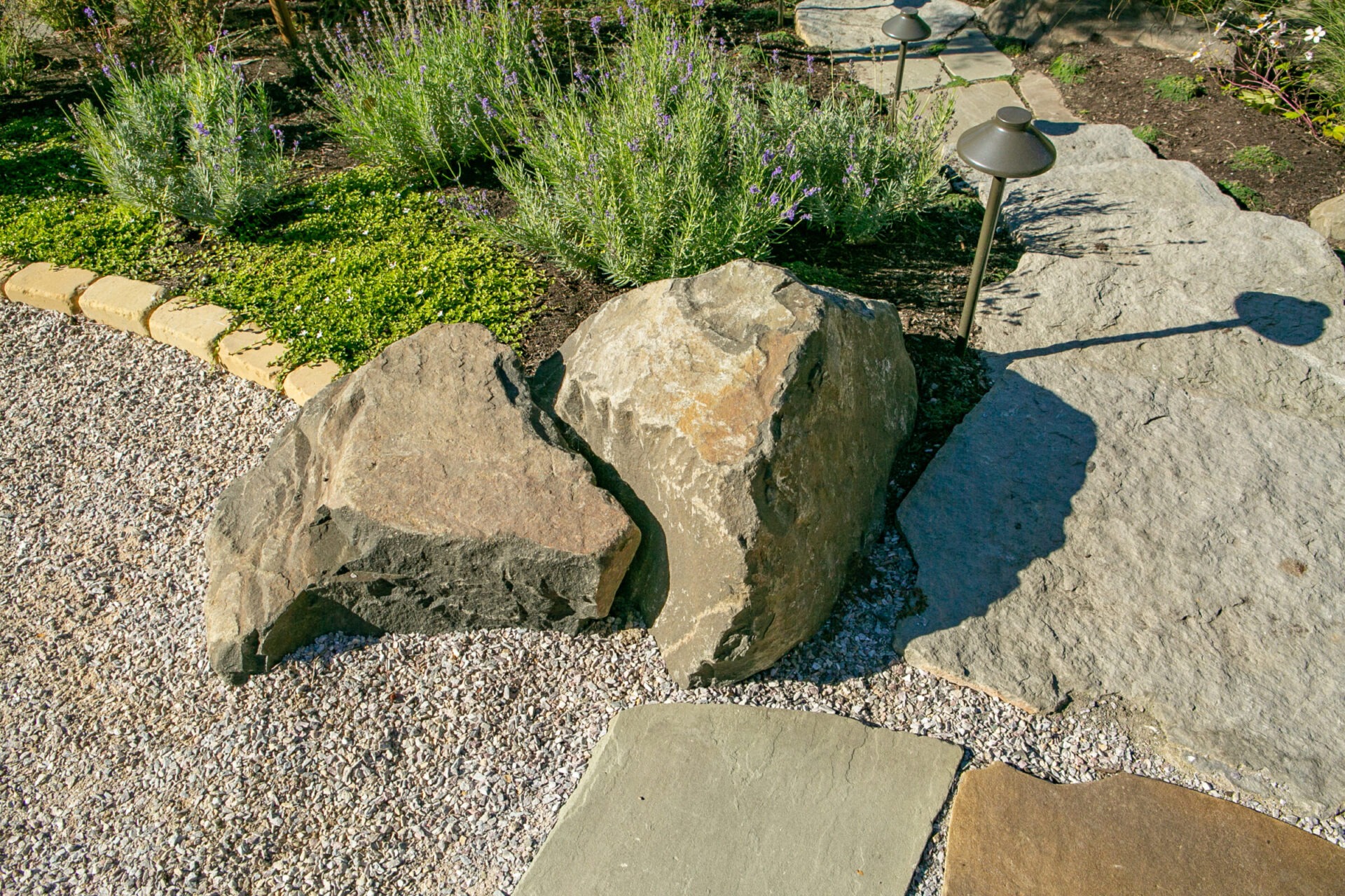 A garden scene featuring large rocks, gravel path, and green plants. A small outdoor light casts a shadow on the stone surface.