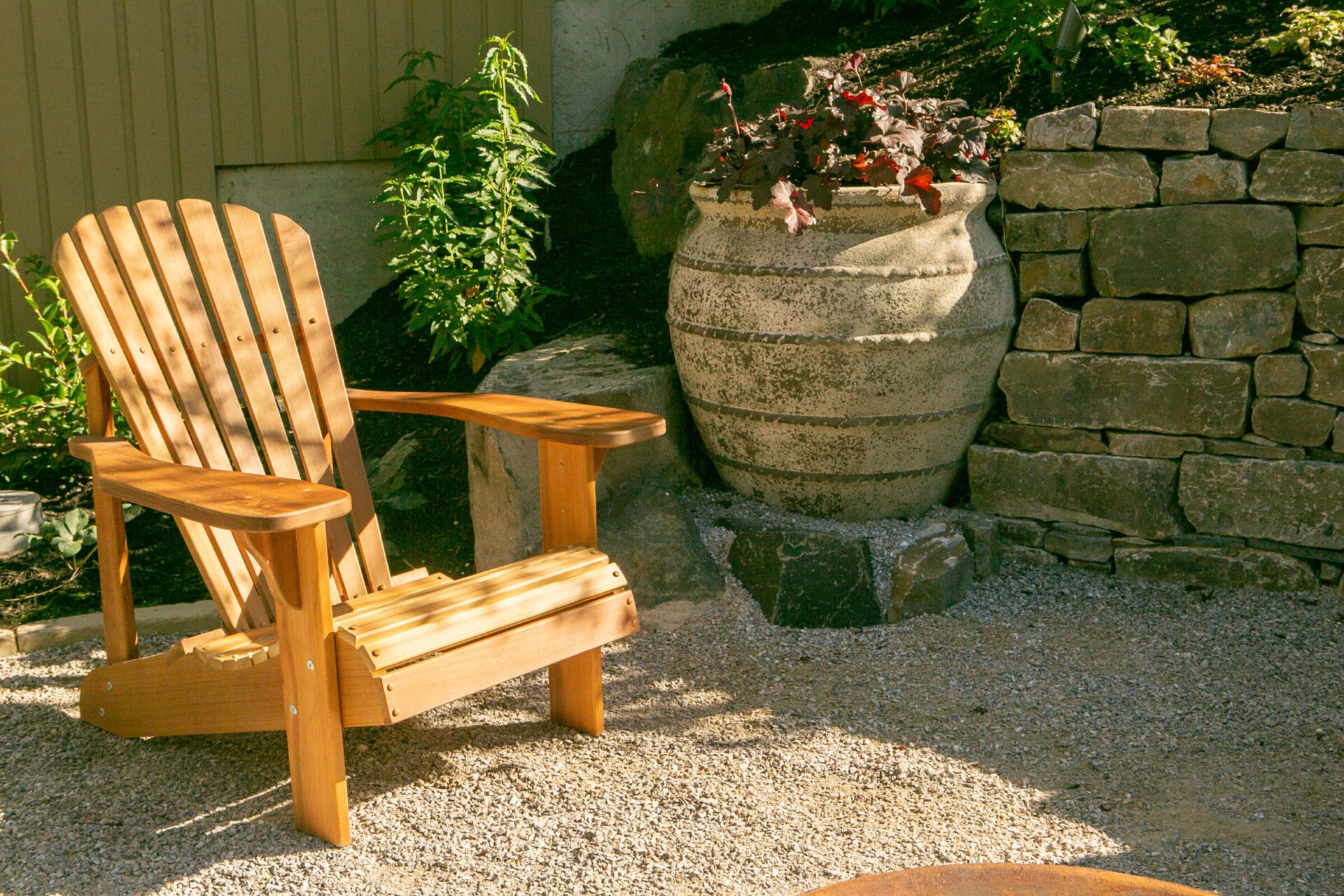 A wooden chair is placed on a gravel surface beside a large decorative pot and stone wall in a garden setting.