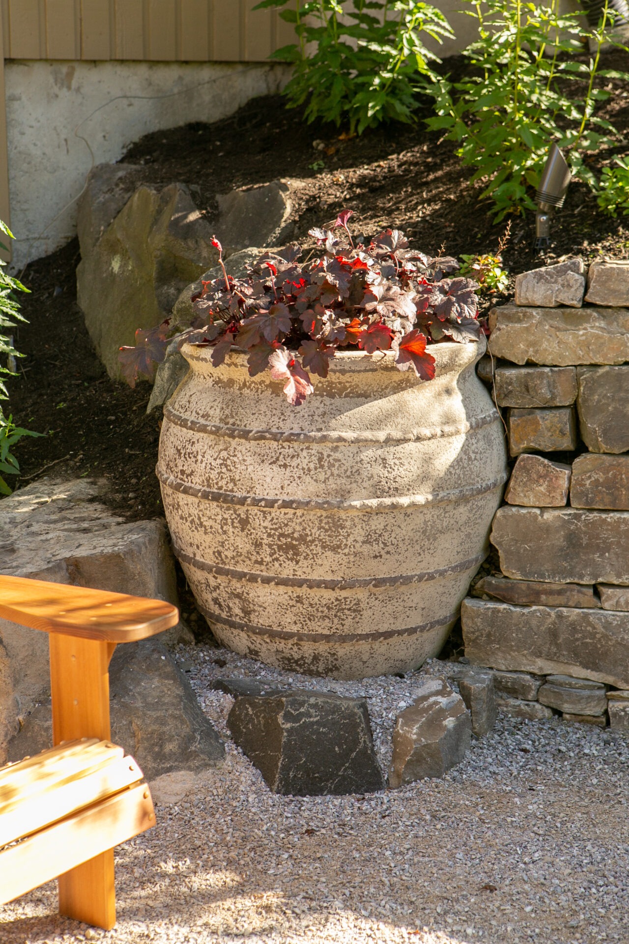 Large stone planter with red-purple foliage, set in a garden beside stacked stone walls and a wooden bench, surrounded by green shrubs.