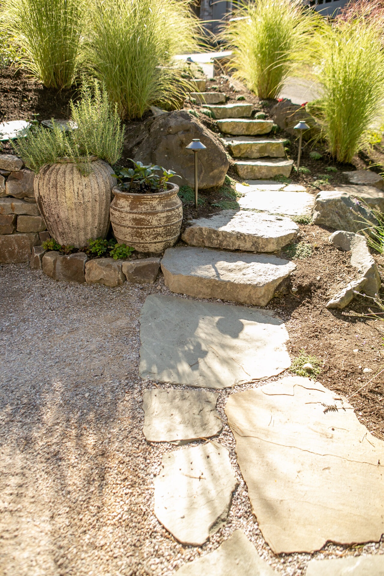 Stone pathway with ascending steps, large planters, ornamental grasses, and well-placed lights create a serene garden atmosphere in bright daylight.