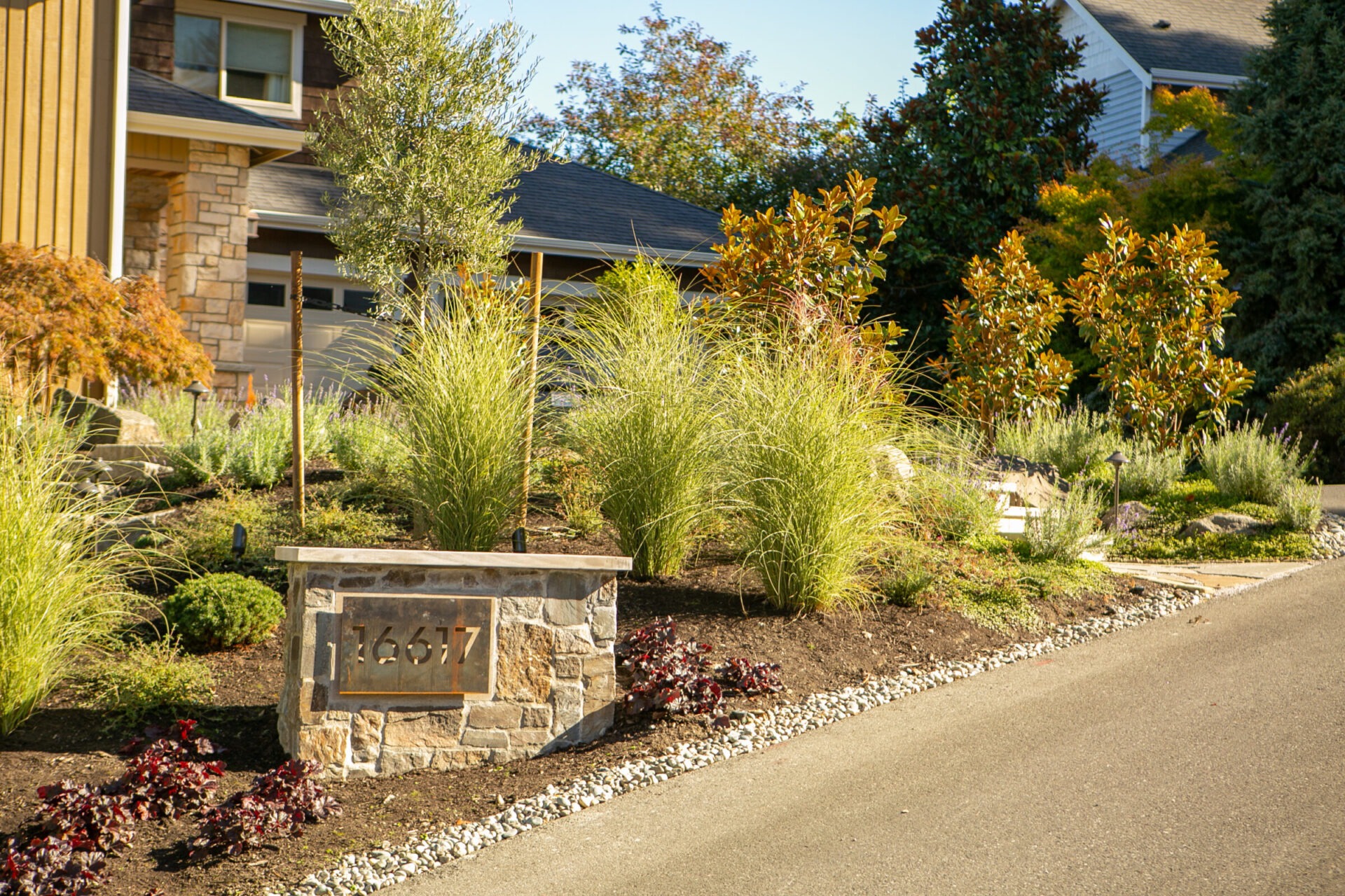 Residential house with modern landscaping, featuring various plants and a stone address marker. The driveway slopes upwards alongside the well-maintained garden.