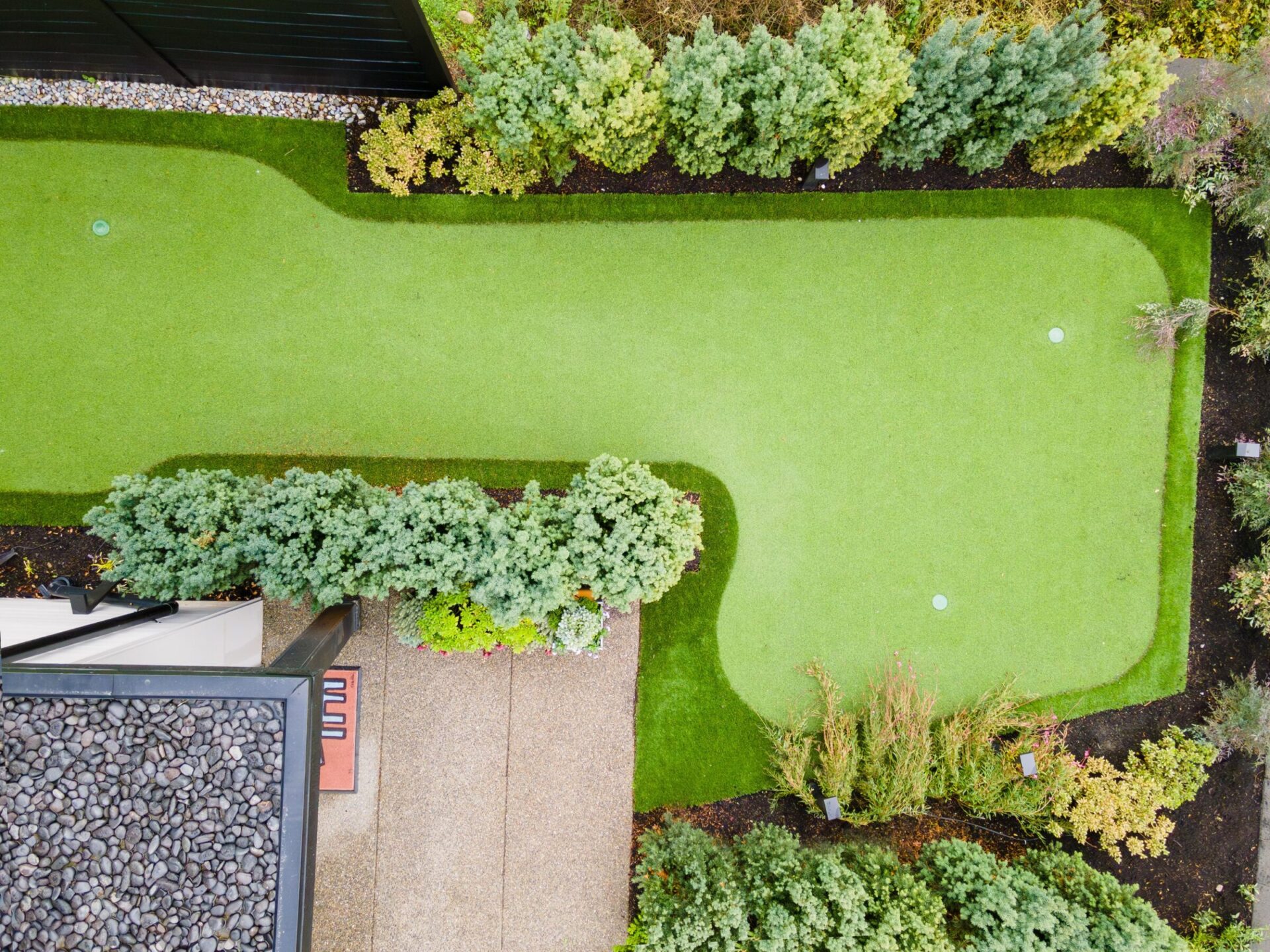 Aerial view of a well-maintained backyard with a small golf putting green, surrounded by lush greenery and decorative shrubs. No people visible.