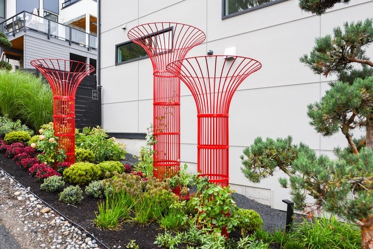 Modern garden with three red metal sculptures, surrounded by neatly arranged plants and shrubs. Contemporary building facade in the background.