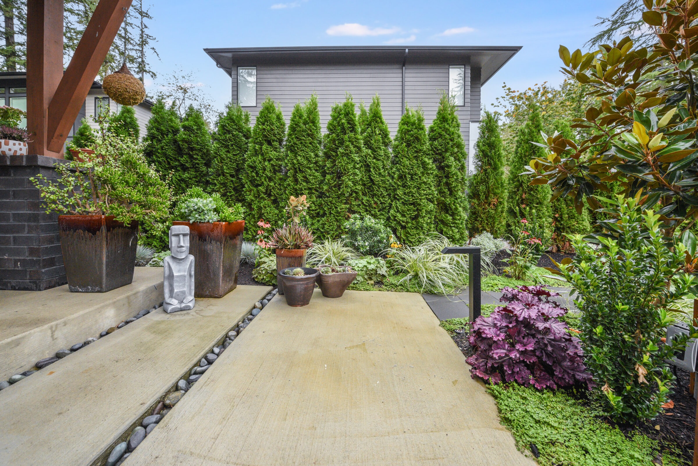 Modern garden with lush greenery and potted plants, featuring a small sculpture and a contemporary gray house in the background under a clear sky.